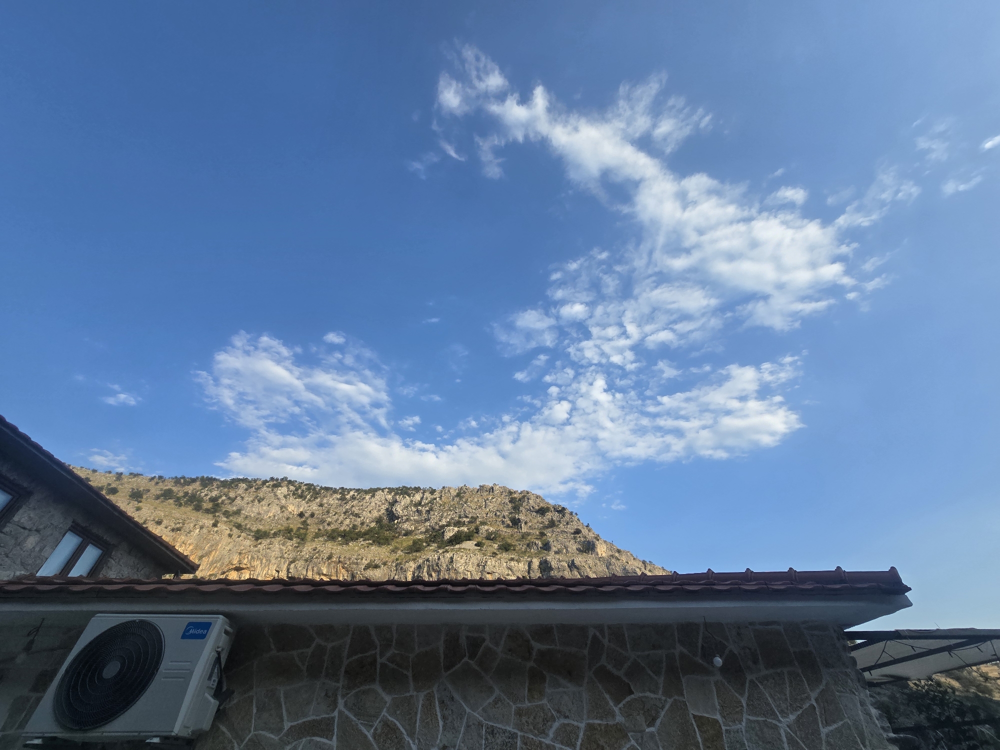 The image depicts a mountain landscape under a bright blue sky with scattered white clouds. In the foreground, the roofline of a building is visible, constructed with reddish-brown tiles and stone walls. An outdoor air conditioning unit is mounted on the stone wall to the left. A portion of another building with a stone facade and a dark-tiled roof is also visible on the far left. The background features a rugged, rocky mountain with sparse vegetation. The scene appears to be a residential area in or near the city of Podgorica, Montenegro, with the mountains characteristic of the region dominating the background. There are no people or animals present in the image.
