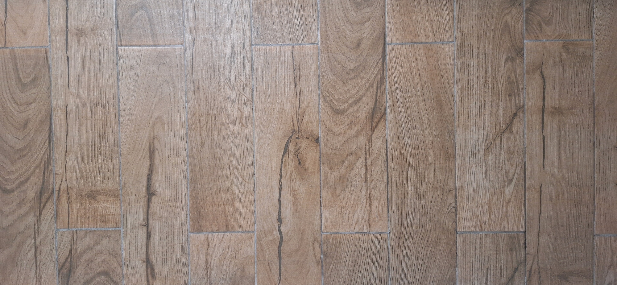 The image displays a close-up, top-down view of a tiled floor designed to mimic the appearance of weathered wood planks. The planks are laid horizontally across the frame and vary slightly in color, ranging from a light tan to a medium brown, with darker brown and grey grain patterns and occasional darker lines suggesting cracks or imperfections in the wood. The grout lines between the planks are thin and a light grey color. The overall texture appears matte, and the lighting suggests an indoor environment. There are no people, animals, or other objects visible in the image, and no specific location in Bila Tserkva, Ukraine, can be identified from the image alone.