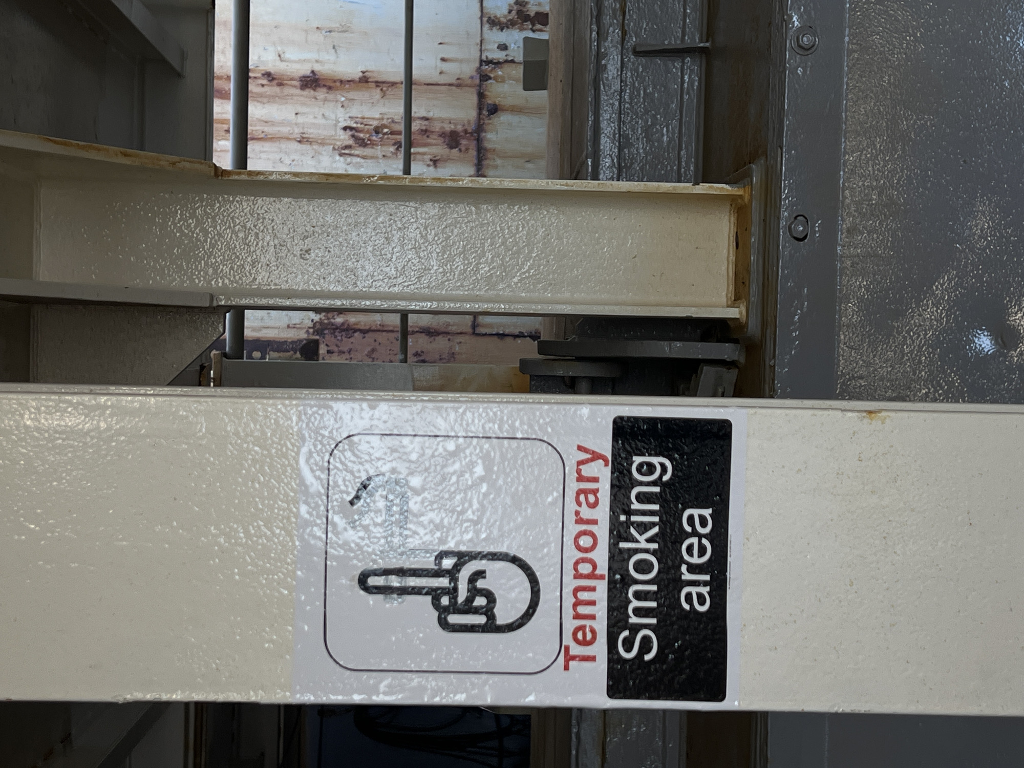 A sign indicating a temporary smoking area is affixed to a painted metal support column in an industrial setting. The sign features a graphic of a hand with the middle finger extended, above the text "Temporary Smoking area". The background shows rusted metal structures and railings, suggesting an outdoor or semi-outdoor environment. The overall scene is industrial and weathered.