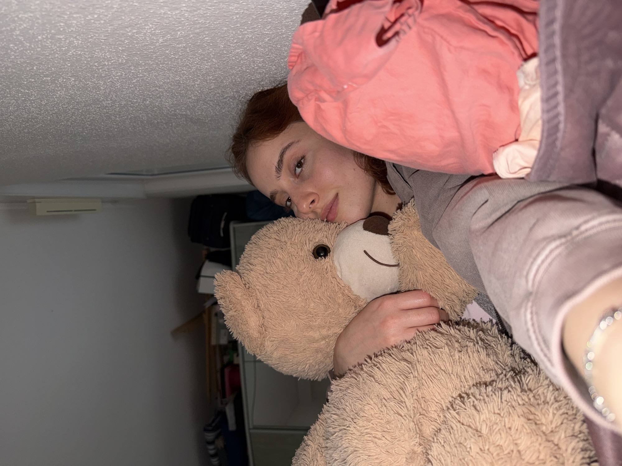 The image displays a young individual, appearing to be a female, lying down or positioned comfortably, gazing directly at the camera. The photo is rotated 90 degrees clockwise, creating an intimate, low-angle perspective. She has reddish-brown hair and a calm, relaxed expression with a subtle smile.
She is wearing a grey top, possibly a hoodie, and is embracing a large, light brown, fluffy teddy bear with dark eyes and a stitched smile. Her left hand rests on the bear's fur, while her right arm, adorned with a silver-colored beaded bracelet, is partially visible on the right side of the frame, contributing to the close-up, personal feel.
The background features a textured white ceiling (likely a popcorn ceiling) at the top of the rotated image, with a white rectangular vent or fixture visible on an adjacent wall. Towards the bottom and left, an indoor room is suggested with dimly lit walls and vague outlines of furniture, shelves, or personal belongings. Blurred pink and purple/grey fabrics, possibly clothing, occupy the immediate foreground on the right.
The scene conveys a moment of comfort and repose, highlighting the bond with the stuffed animal. The lighting is soft and even, typical of indoor artificial light. No specific time of day is discernible. There is no visible text in the image. The setting is an indoor, private space, situated in Strasbourg, France, though no direct visual cues in the image confirm this location.