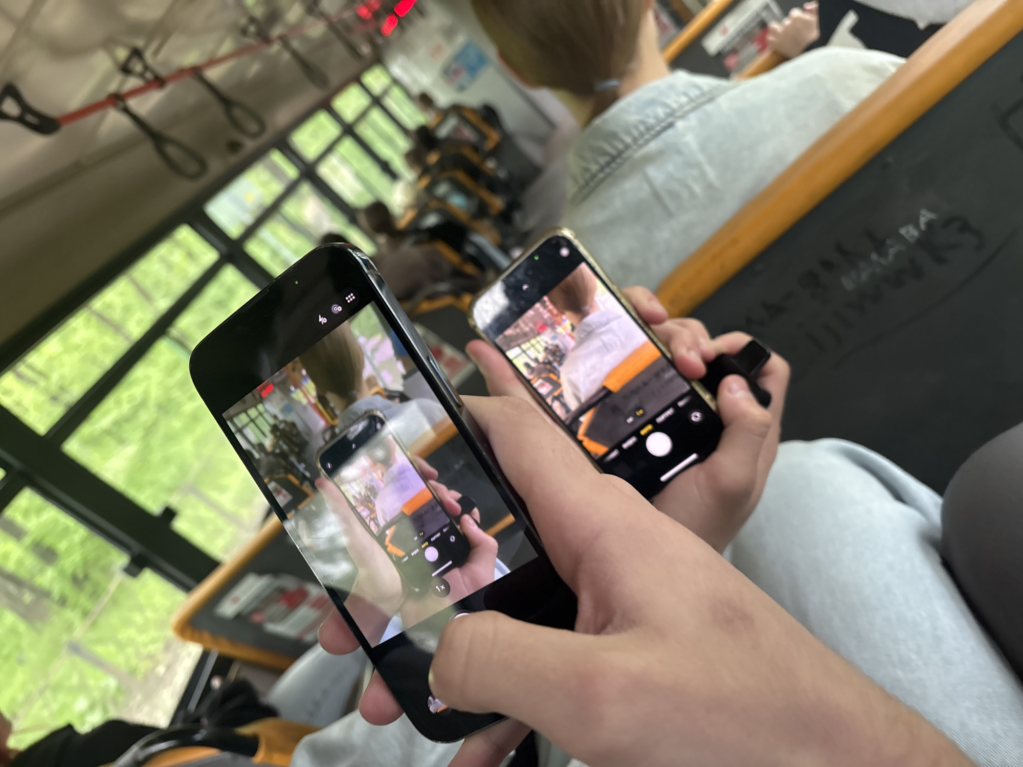 The image captures a unique recursive photographic effect, commonly known as the Droste effect, set within what appears to be the interior of a public transport vehicle. In the foreground, two hands hold two smartphones. The phone on the left, a black model, displays its camera interface and is aimed at the phone on the right. The gold-colored phone on the right also displays its camera interface, which shows a live feed or reflection of the black phone's screen, creating an infinite loop of phones within phones. The hands appear to be actively engaging with the devices, likely taking a photo or video of this recursive view.
The setting is a bus or similar public transport vehicle, evidenced by the characteristic overhead red strap handles with black loops and large windows that reveal lush green foliage outside, indicating it is daytime. Rows of seats can be seen in the background, with blurred figures occupying some of them. A person with light-colored hair, wearing a light denim-like shirt, is partially visible from behind in the mid-ground. On the back of a seat or surface in front of this person, faint text or graffiti can be seen, which includes characters resembling "АА-ДЛАБА" and "ЦЛVWK3". The lighting is bright and natural, suggesting good daytime visibility.
