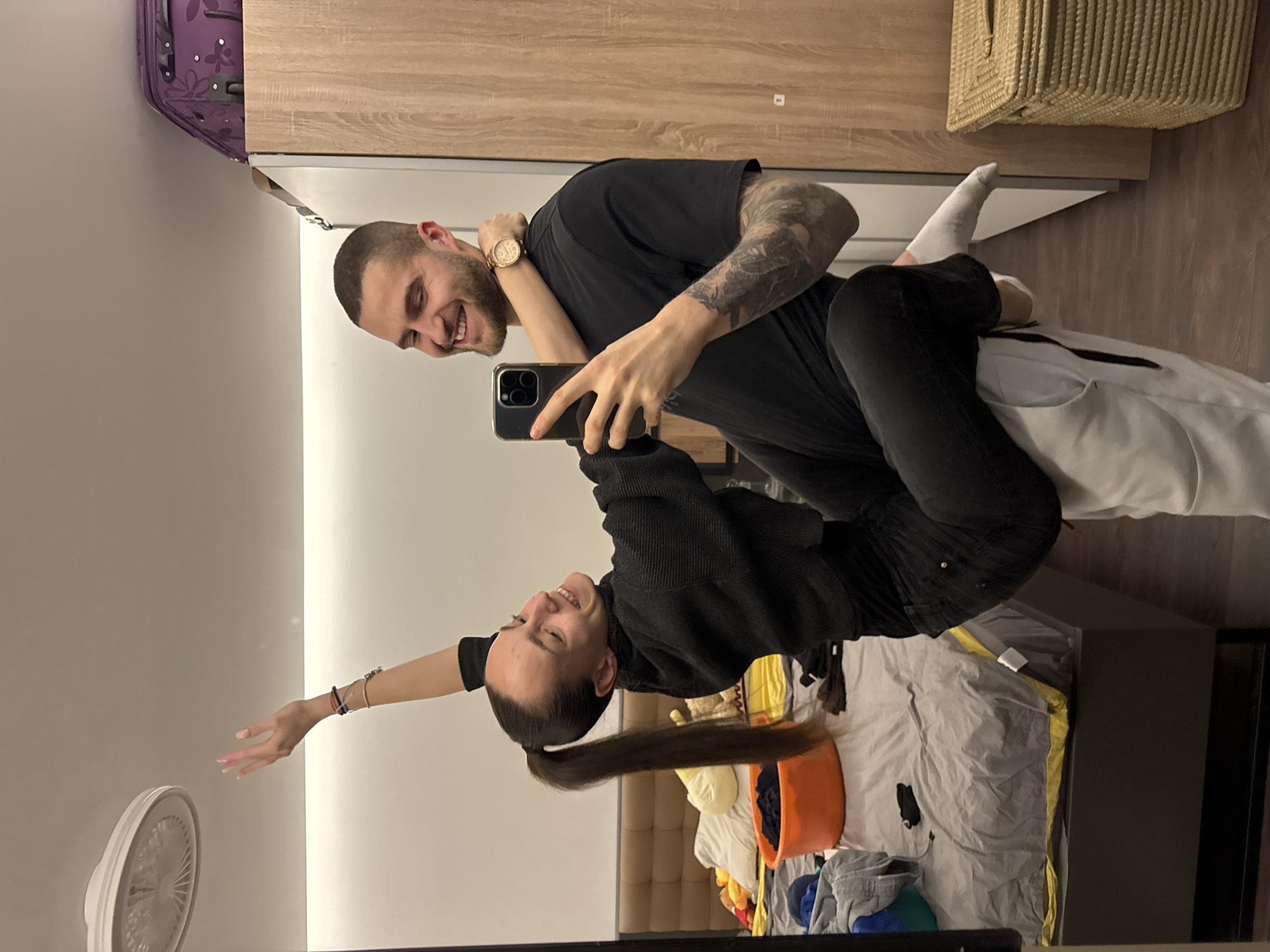 The image is a mirror selfie depicting a young adult female and male engaged in a playful interaction within what appears to be a private residence or hotel room. The female, with dark hair tied in a high ponytail, wears a black long-sleeved top and black pants. She is holding a dark-colored smartphone with multiple lenses, angled to capture their reflection. Her left arm is wrapped around the male's neck, and her right arm is extended upwards with fingers splayed. She is smiling broadly and looking towards the male. She appears to be partially seated on the male's upper left leg/hip.
The male, with short dark hair and a beard, is wearing a black t-shirt, light-colored pants, and white socks. His left arm is around the female's waist, supporting her. He displays tattoos on his left forearm and wears a gold-colored wristwatch on his left wrist. He is smiling broadly and looking towards the female.
The background, a reflection in the mirror, reveals details of the room. To the left, a bed with a dark grey duvet and a yellow fitted sheet is visible, upon which various items of clothing are scattered, along with an orange plastic tub containing more clothes. A padded, brown headboard is positioned behind the bed. Above, a white ceiling features a round, white light fixture, possibly a fan. To the right, a light-colored wooden wardrobe or cabinet stands, topped with a purple patterned suitcase. A woven, light-colored basket is on the dark wooden floor next to the wardrobe. The image itself contains no identifiable landmarks or specific characteristics to determine a precise location within Sofia, Bulgaria; it depicts a generic interior space.