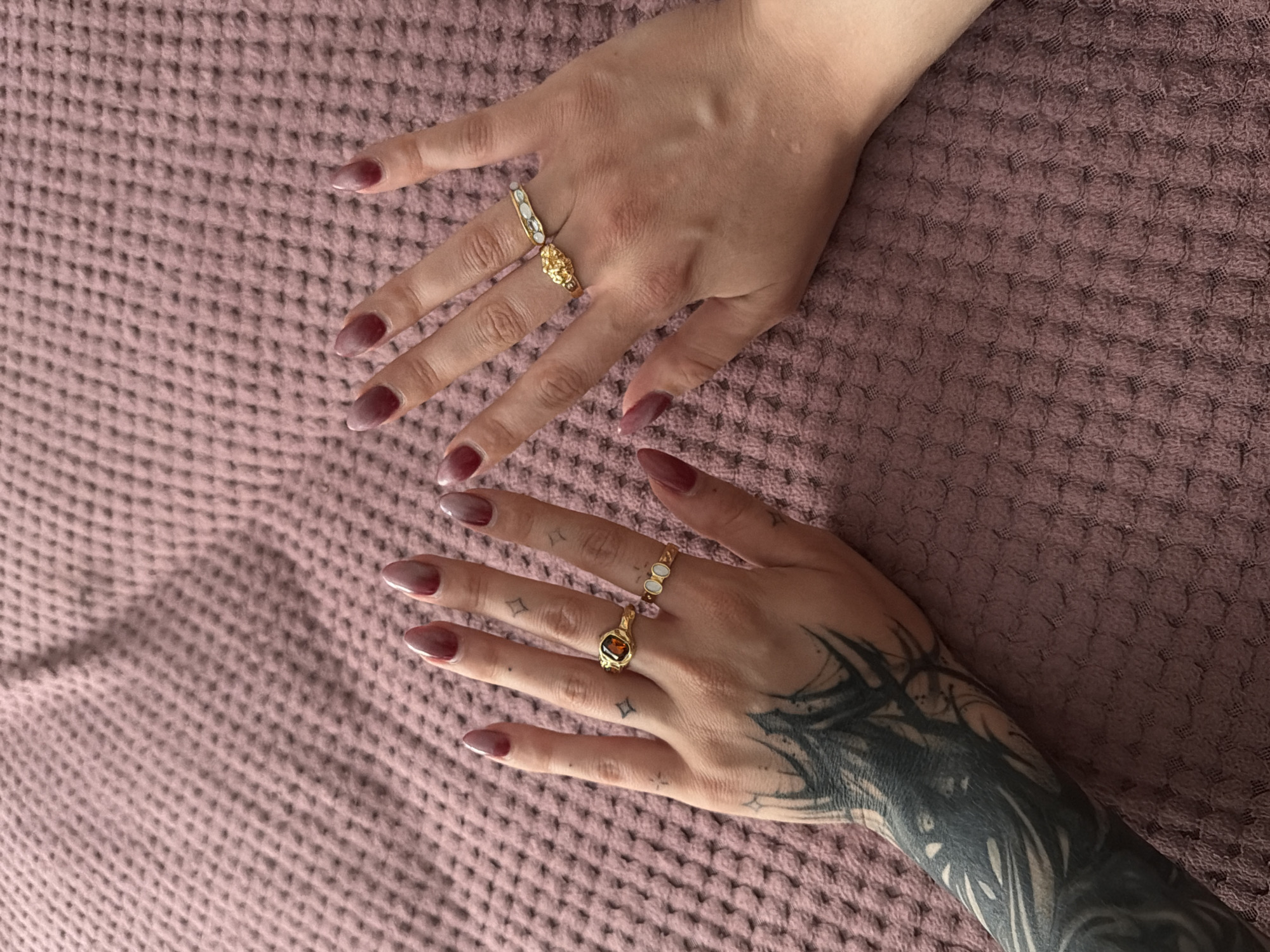 Two hands, adorned with dark pink gradient manicured fingernails and gold rings, rest on a textured pink surface. The left hand features a gold ring with a dark orange stone and a smaller gold ring with three oval, white stones. The right hand displays a decorative gold ring with a textured, floral-like design. The left forearm is heavily tattooed with black ink, depicting intricate, dark designs. The background consists of a pink, textured fabric, possibly a blanket or bedding. The lighting is soft, casting subtle shadows. The image appears to be a close-up shot, focusing on the hands and rings.