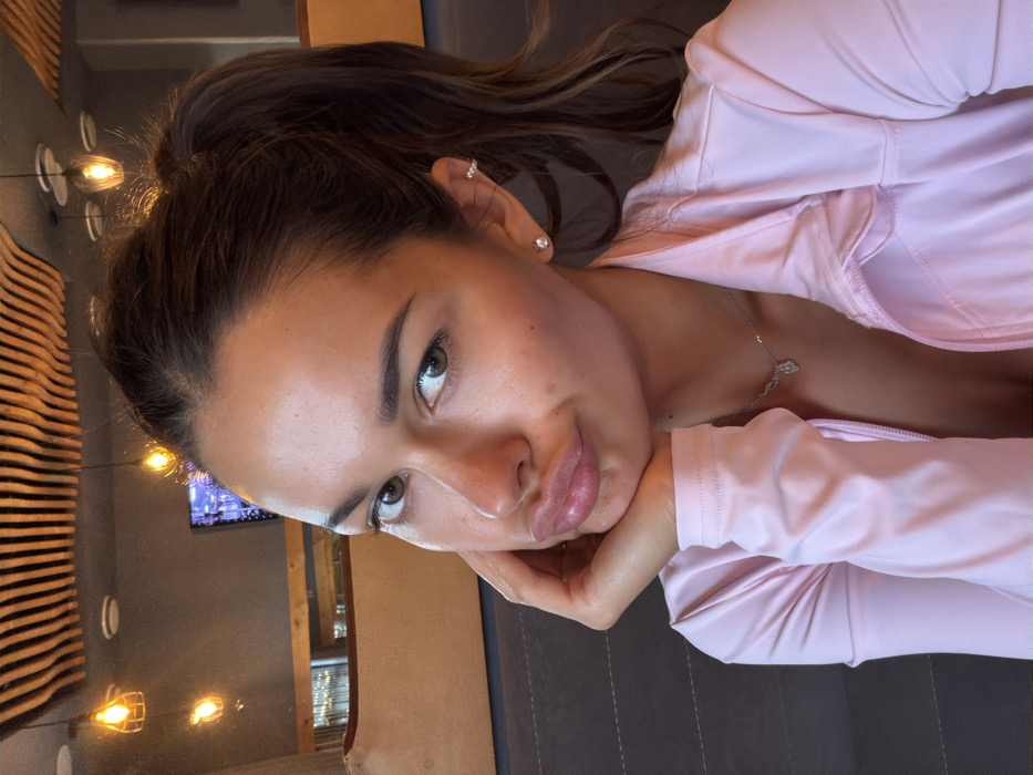A young adult female is depicted in a close-up, chest-up portrait within a commercial establishment in Krasnoyarsk, Russia. The subject has dark brown hair pulled into a high ponytail and appears to have a tanned complexion. She looks directly at the camera with her lips pursed in a "duck face" expression. Her right hand rests with fingers supporting her chin. She wears a light pink, long-sleeved zippered top, partially unzipped, revealing a glimpse of undergarment. A silver necklace with a small pendant adorns her neck. Her right ear features a small round stud earring in the lobe and a small cuff or hoop earring on the upper helix.
The background reveals the interior of what appears to be a restaurant, cafe, or lounge. The ceiling exhibits a distinctive design composed of parallel, undulating wooden slats. Several exposed filament light bulbs in black wire cage fixtures hang, providing warm, ambient illumination. A dark gray upholstered booth or bench is visible in the foreground, with lighter-colored upholstered seating further in the background. A large flat-screen television displaying blue and purple graphics is mounted on a far wall.
