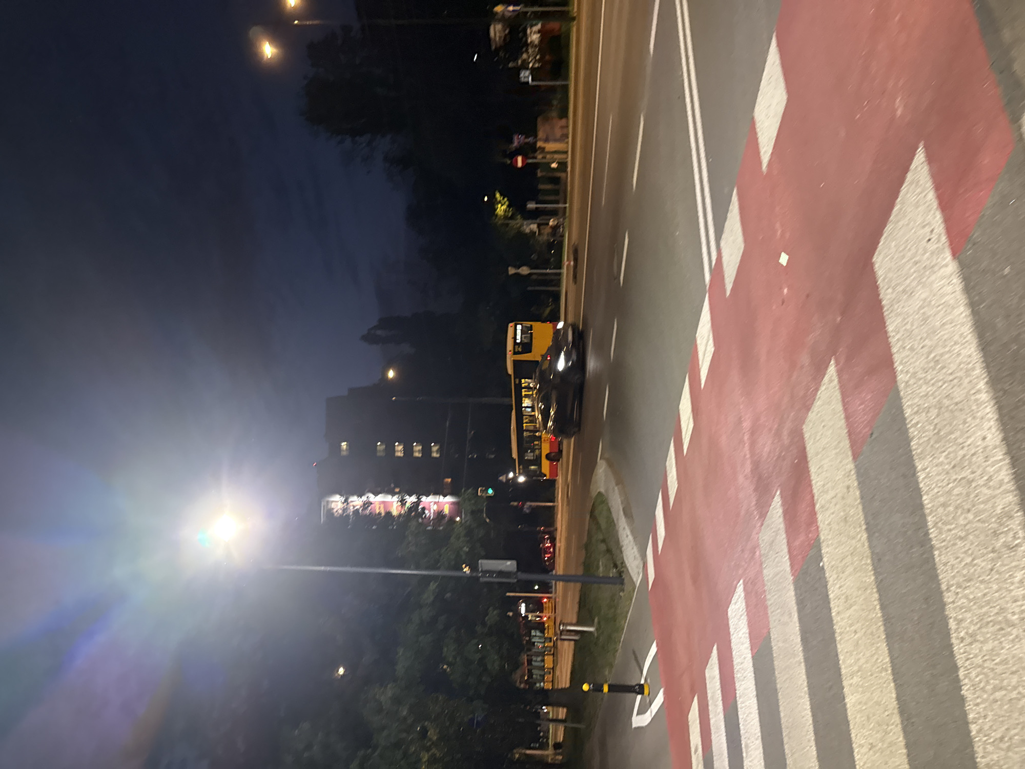 An eye-level, medium shot captures a street scene at night in an unknown city. The foreground is dominated by a red and white striped pedestrian crossing. Beyond this, a paved road with white lane markings stretches into the distance. A dark-colored sports car is in motion on the road, to the right of center. Further down the road, a yellow and red bus is visible, also in motion. Several streetlights illuminate the scene, casting bright glows that cause lens flare in the upper left quadrant of the image. Tall, dark buildings are visible in the background, some with illuminated windows. Trees line the left side of the road. The sky is dark blue with some faint clouds.