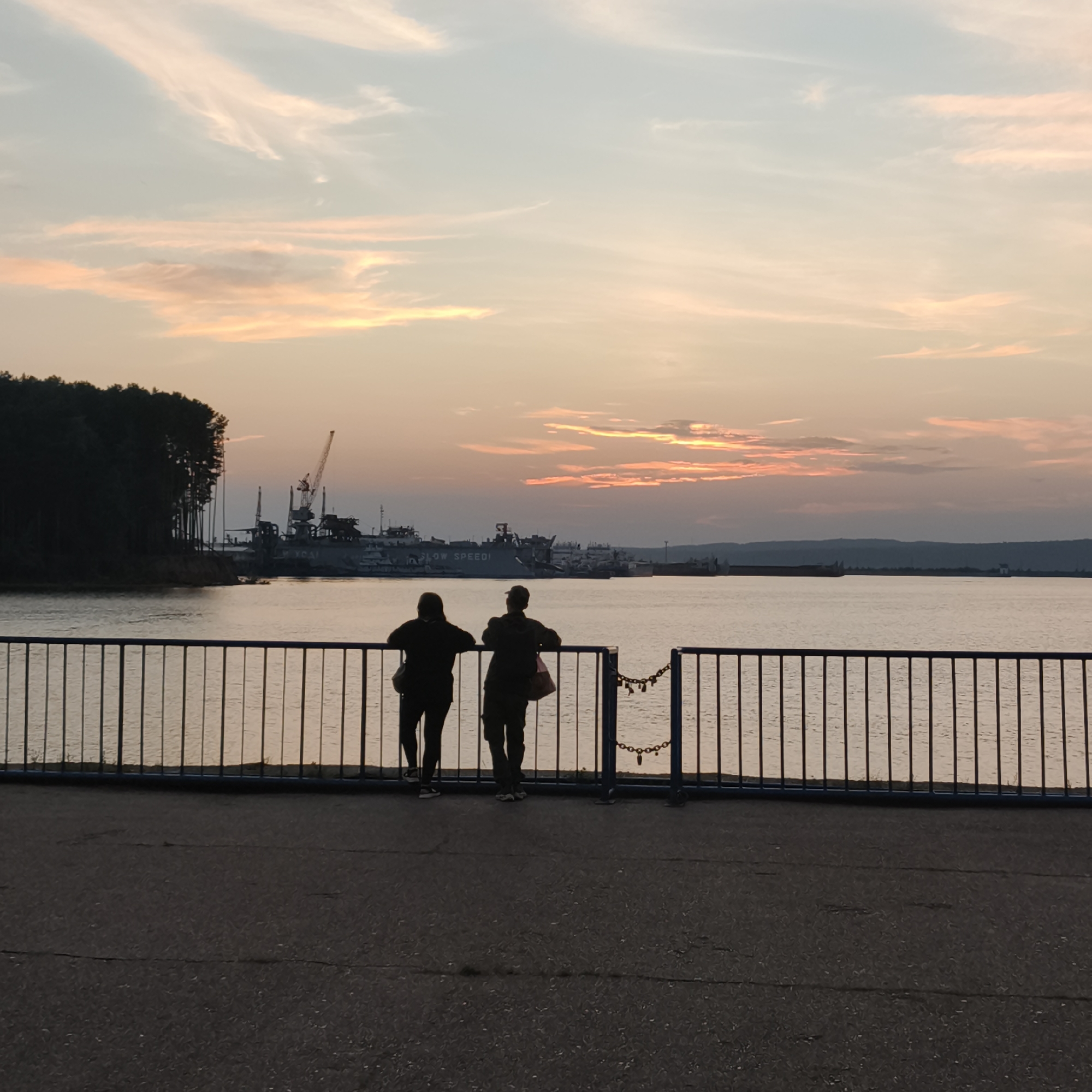 Two figures are silhouetted against a sunset sky as they stand by a black metal fence overlooking a body of water. In the distance, industrial structures including cranes and ships are visible on the far bank, partially obscured by trees along the shoreline. The sky is painted with soft hues of pink, orange, and blue, with wispy clouds. The foreground consists of a paved area. This scene is captured on the embankment of the Kama River in Chaikovsky, Russia.