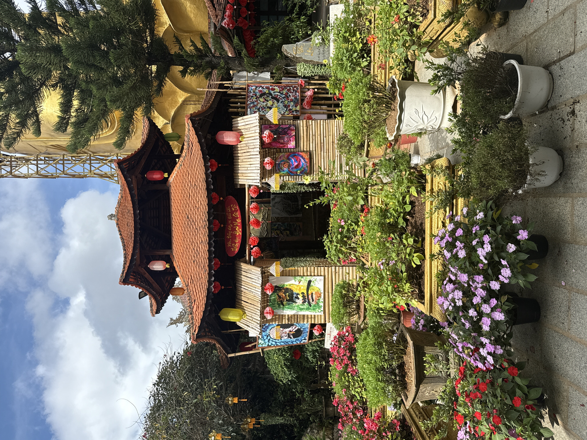 The image depicts the exterior of a building with traditional East Asian architecture, likely a temple or shrine, situated in Lâm Viên - Đà Lạt, Vietnam. The building features multiple tiered roofs with orange tiles, ornate eaves, and is adorned with red lanterns. The walls of the structure are constructed from bamboo, and several colorful paintings are displayed on the facade.
In the foreground, a vibrant garden is meticulously arranged with numerous potted plants. These include lush green bushes, bright red flowers, and delicate purple blooms. Various sizes and styles of planters, some made of stone and others of ceramic, are interspersed throughout the scene.
Behind the main building, a massive, golden structure, possibly a statue or a portion of a larger complex, is partially visible, along with the skeletal framework of scaffolding. A tall pine tree stands to the right, its branches extending towards the golden structure. The sky above is a clear blue, dotted with scattered white clouds. The overall impression is one of a tranquil and culturally rich environment, set within a botanical garden or spiritual site. No people are present in the image.