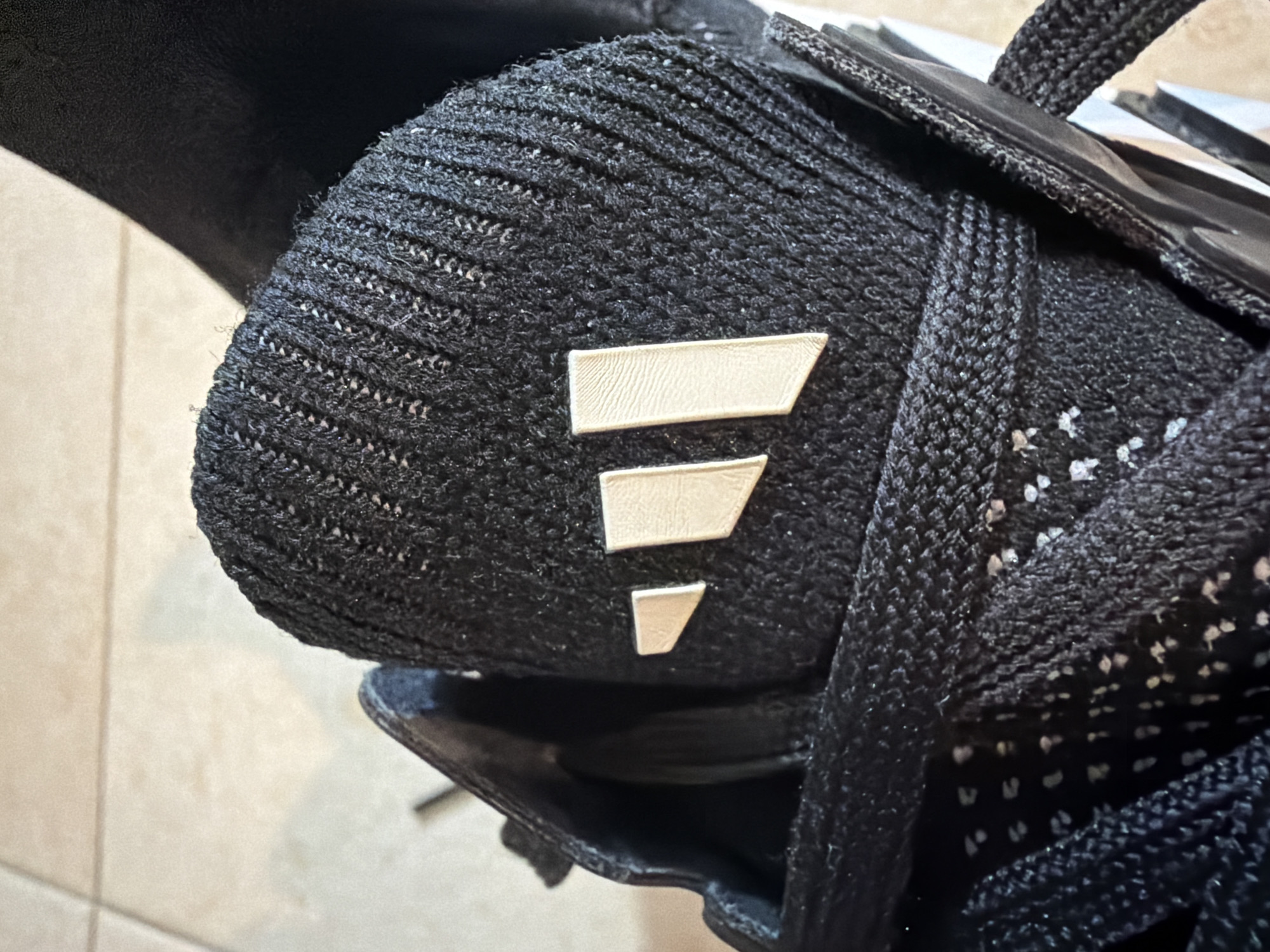 The image shows a close-up of a black Adidas athletic shoe, specifically the top and tongue area. Three light-colored, rectangular stripes, a hallmark of the Adidas logo, are prominently displayed on the textured black fabric of the shoe's tongue. The shoe is laced with thick black laces. The background consists of a light-colored tiled floor, suggesting an indoor setting. The image is a detailed shot of the shoe's branding and material. The location is not discernible from the image itself.