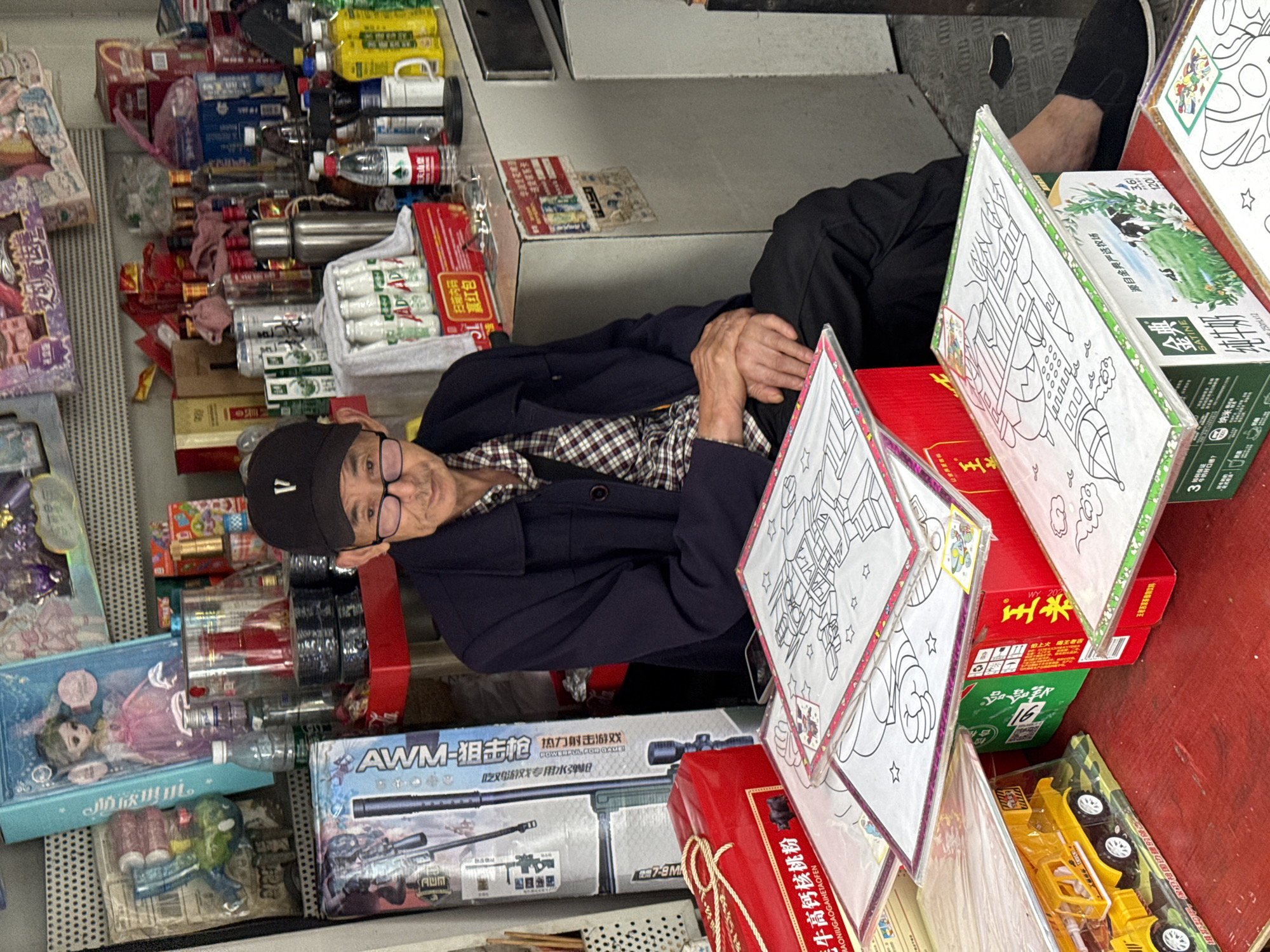 An elderly Chinese man is resting in what appears to be a small shop or stall, possibly in Jiangjin, China. He is seated with his hands clasped together. He is wearing glasses and a dark cap. The surrounding space is filled with various goods for sale, including bottled drinks, toys, boxes of what appears to be food or household products, and packaged items. The items are densely packed, filling the shelves and surrounding areas. There are toys, including a toy gun, and some color-in posters. The scene is indicative of a small business operation.