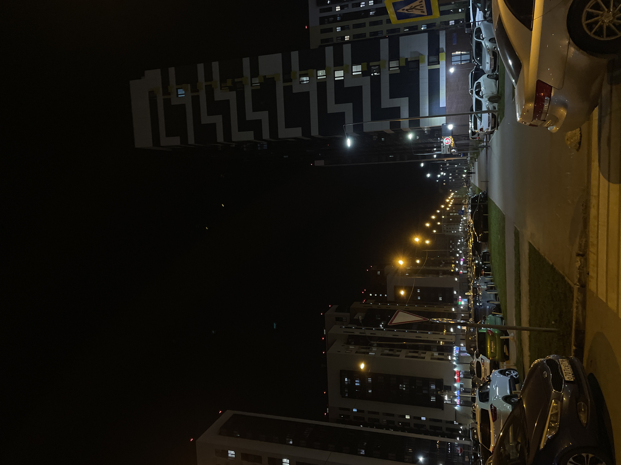 The image depicts a nighttime street scene in Kazan, Russia. The foreground includes a parked silver car and a crosswalk. A line of parked cars extends down the street, illuminated by streetlights. On either side of the street are tall, modern apartment buildings. One building has a distinctive black and white zigzag pattern on its facade. There are several traffic signs along the street.