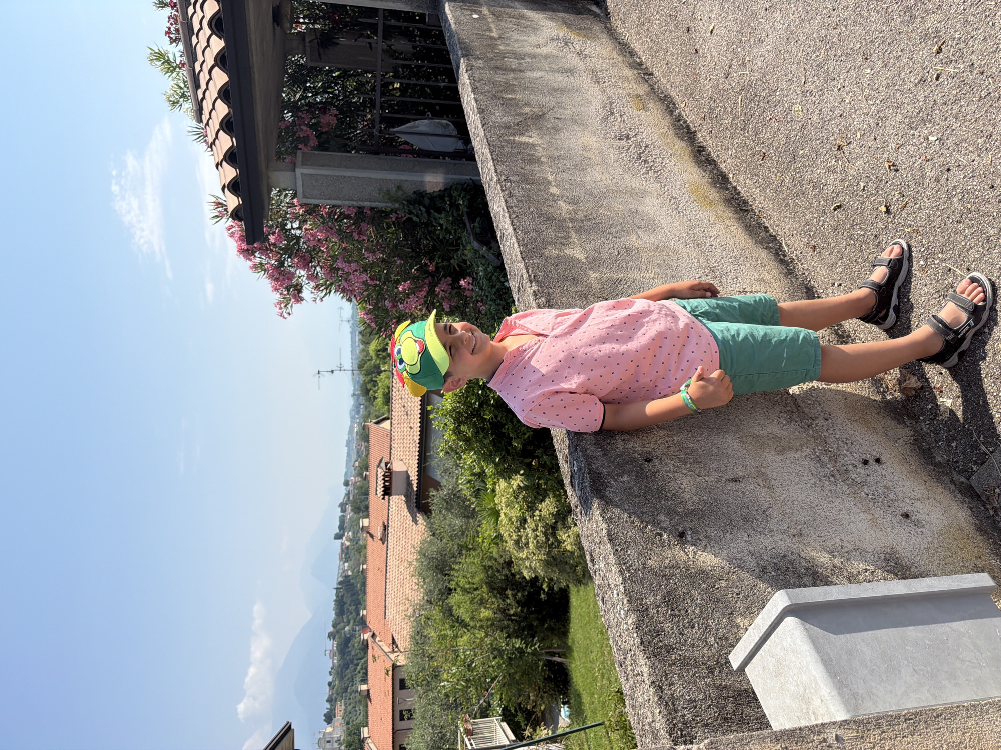 A young boy wearing a pink polka-dot shirt, green shorts, and a green baseball cap with a cartoon character on it is standing by a concrete wall. He is smiling and looking towards the viewer. The background shows a hilly landscape with houses, trees, and mountains in the distance. The scene is set on a sunny day. The location is identified as Polpenazze del Garda, Italy, likely on a residential street with a view of the surrounding countryside. The concrete wall appears to be a retaining wall or the edge of a pathway. A grey metal post is visible on the left side of the frame, attached to the wall.