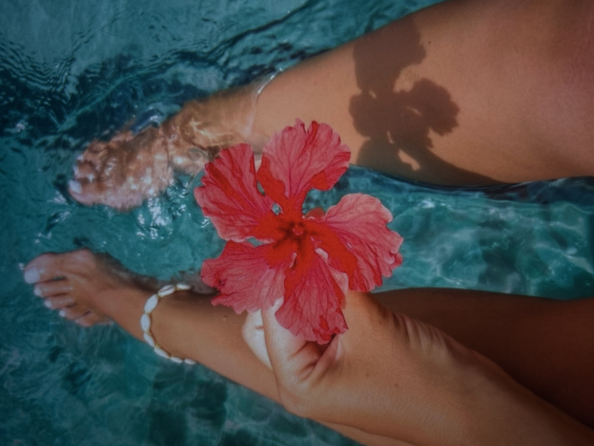 A first-person perspective shot focuses on a woman's legs submerged in clear turquoise water. Her right foot, adorned with a shell anklet, is visible near the top left of the frame. She holds a vibrant pink hibiscus flower in her right hand, positioned in the center of the image, with her fingers delicately grasping its stem. Her left leg extends towards the bottom right, partially submerged and casting a soft shadow on the water's surface. The water's movement creates ripples and reflections, highlighting the clarity and color of the pool. The scene evokes a sense of relaxation and tropical serenity.