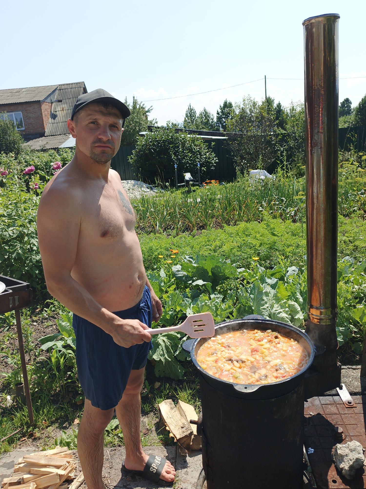 The image shows a man standing in front of a large cauldron of food being cooked outdoors. The man is wearing blue shorts and a black baseball cap, and he is shirtless. He is holding a pink spatula and appears to be stirring the food in the cauldron. The cauldron is placed on a metal stand, and there is a tall metal chimney next to it. The scene is set in a garden or backyard with lush green plants and a brick house in the background. There are also piles of firewood on the ground, suggesting an outdoor cooking event. The sky is clear and blue. The location is identified as Medvedyovka, Russia.