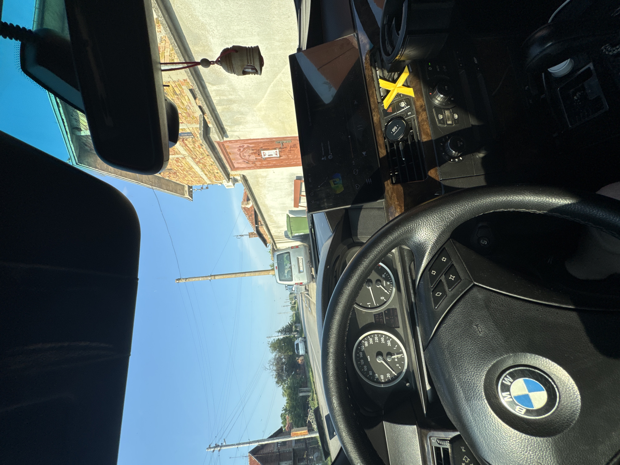 The scene is viewed from inside a vehicle, specifically a BMW (indicated by the steering wheel emblem and dashboard design), from the driver's perspective, looking forward through the windshield. The vehicle appears to be stationary or moving slowly on a paved two-lane road.
**Vehicle Interior:**
The black leather steering wheel with a silver BMW emblem is prominent in the lower left. The instrument cluster shows a speedometer on the left, an RPM gauge on the right, and a digital display between them indicating "12.2 l/100km" and "82987 km". An aftermarket rectangular tablet-style display is mounted on the center of the dashboard, showing a dark interface, likely a music player or navigation system, with various icons and text. Below this screen are climate control vents and knobs. A yellow "X" shaped object is affixed to the right of the central vents. A round black magnetic phone mount is visible on the dashboard to the left of the vents. A small wooden ornament hangs from the rearview mirror by a red string. The sky is visible through the top of the windshield and above the sun visor.
**Exterior Environment:**
The road is asphalt and appears to curve slightly to the right in the distance.
On the immediate right, a brick building with a prominent red, ornate wooden door is visible behind a concrete wall. Above the building, a light blue roof structure is partially visible. A wooden utility pole stands near the curb on the right.
Further down the road on the right, a silver van with Bulgarian license plate **CT 6324 PB** is parked facing away from the camera. Beyond it, green trash bins are visible.
On the left side of the road, another utility pole stands, followed by a residential building, partially obscured by trees and bushes. A white car is parked further down the road on the left.
The sky is clear and blue, indicating daytime with strong sunlight. Overhead power lines crisscross the scene between the utility poles.
**Location:**
Based on the license plate prefix "CT" (Stara Zagora Province) and detailed visual matching of the distinctive brick building with the red door and adjacent concrete wall, the location is identified as **ул. Първа (Părva St.) in Sarnevo, Stara Zagora Province, Bulgaria**, specifically near house number 32, facing generally northwest down the street.