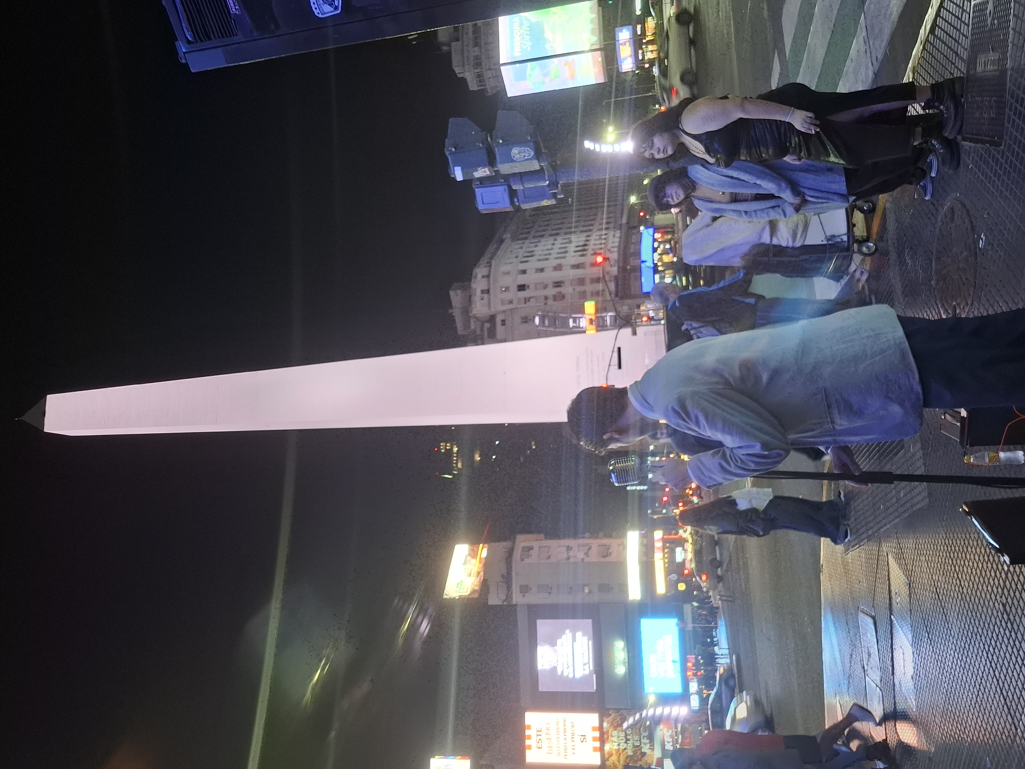 At night, the Obelisco stands tall in the city of Buenos Aires, Argentina, illuminated against the dark sky. A man wearing a light-colored jacket is speaking into a microphone, standing on a metallic grid surface. Nearby, two women are looking in his direction. Advertisements and city lights brighten the surrounding buildings.