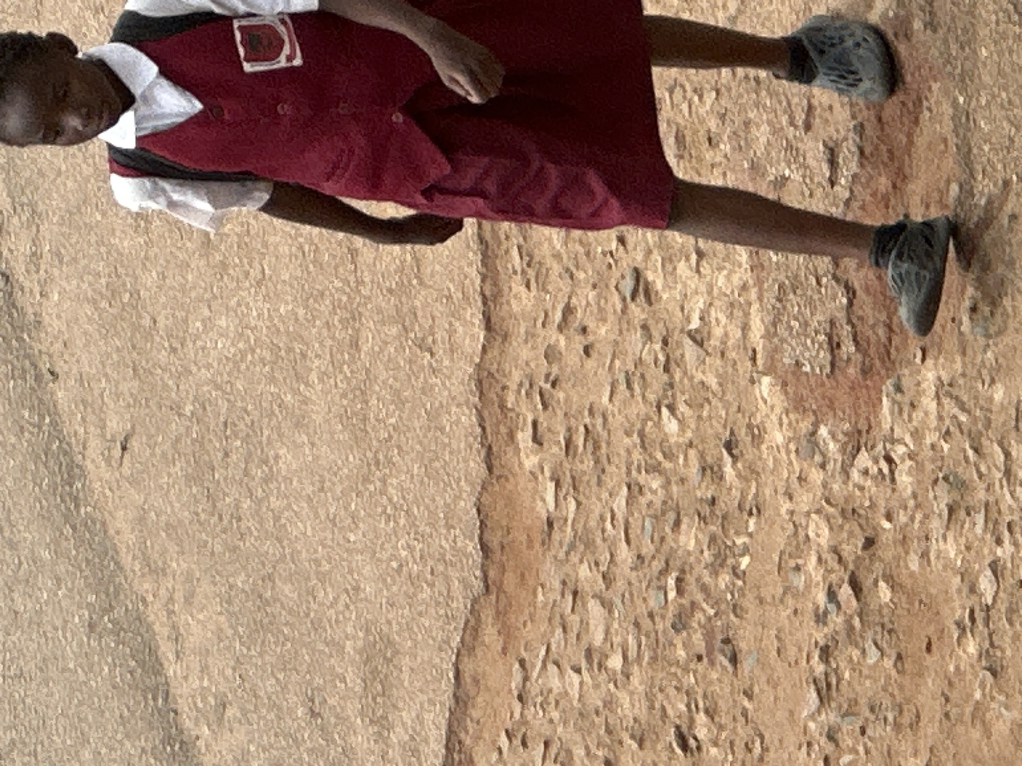 A young girl in a school uniform, consisting of a white collared shirt, a maroon vest, and a maroon skirt, walks across a dirt path. The girl carries a black backpack. She is wearing grey shoes and dark socks. The ground is covered with dirt and small stones. The background is out of focus, but appears to be a dusty, outdoor environment. The scene is likely in Jos, Nigeria, given the dry, reddish soil and the style of the girl's uniform, which is typical for many schools in the region.