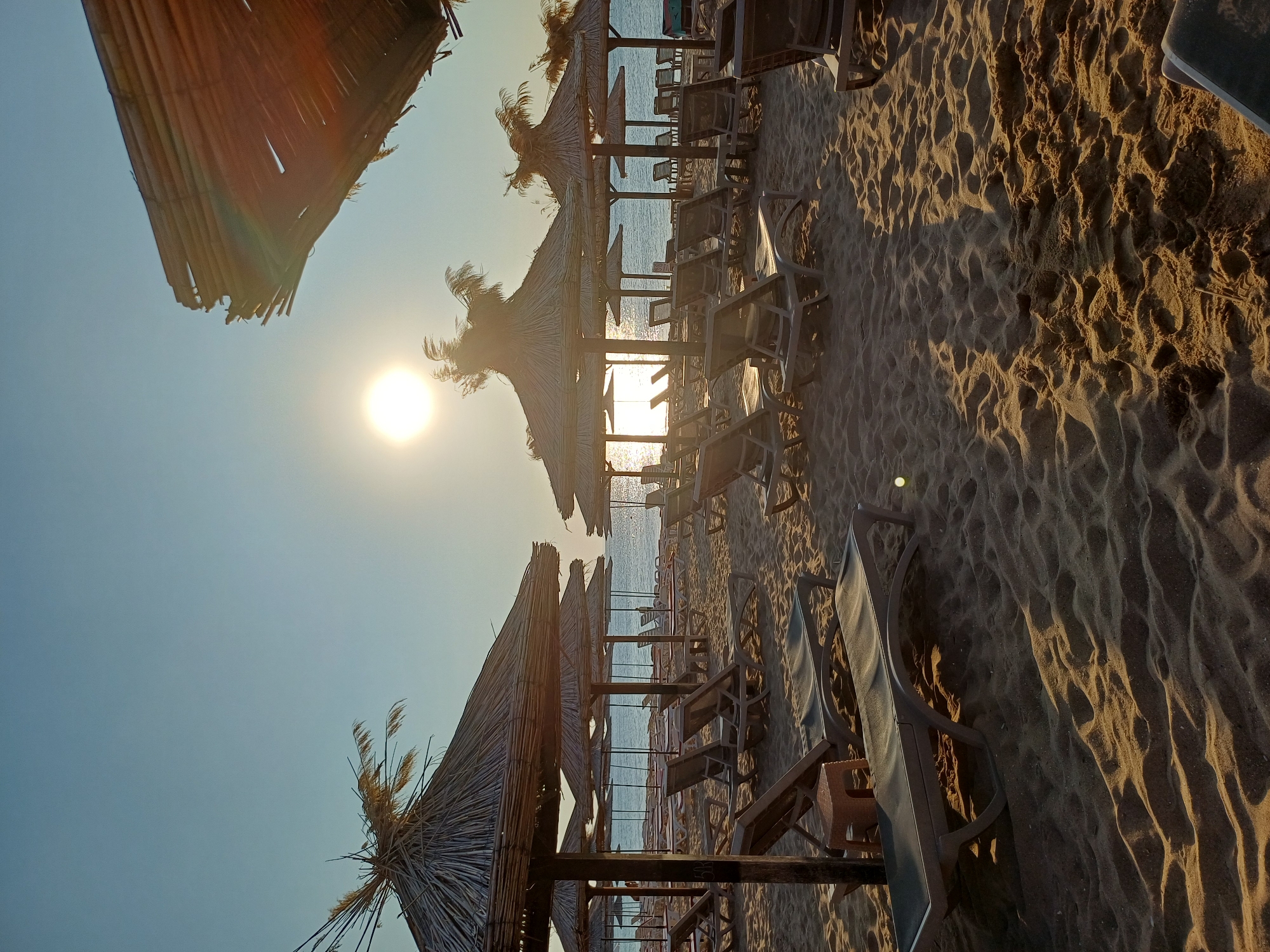 The image depicts a sandy beach at sunrise or sunset, with the sun visible in the upper center of the frame. Numerous empty lounge chairs and thatched umbrellas are arranged on the sand. In the background, the calm sea is visible. The scene appears to be on a resort beach, potentially located in Varna, Bulgaria, given the context. There are no people visible in the frame. The lighting suggests a warm, golden hour.