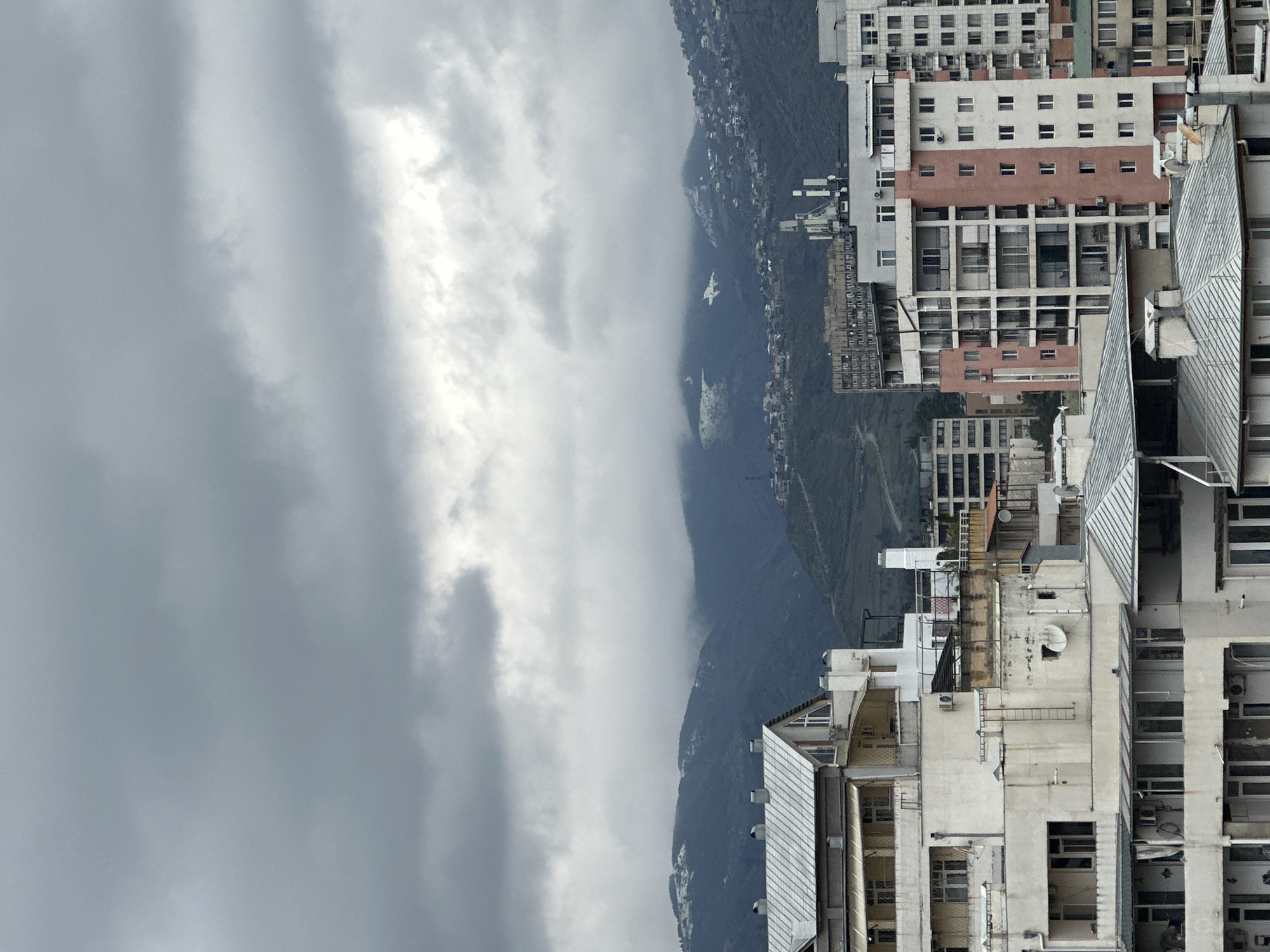 The image shows a view in Tbilisi, Georgia, where city buildings meet a mountain shrouded in clouds. The weather appears overcast. The buildings are a mix of architectural styles, some with light-colored facades and repeating window patterns, others with more modern, possibly metallic, roofs. The mountain rising in the background is densely populated with buildings, partially obscured by low-hanging clouds.