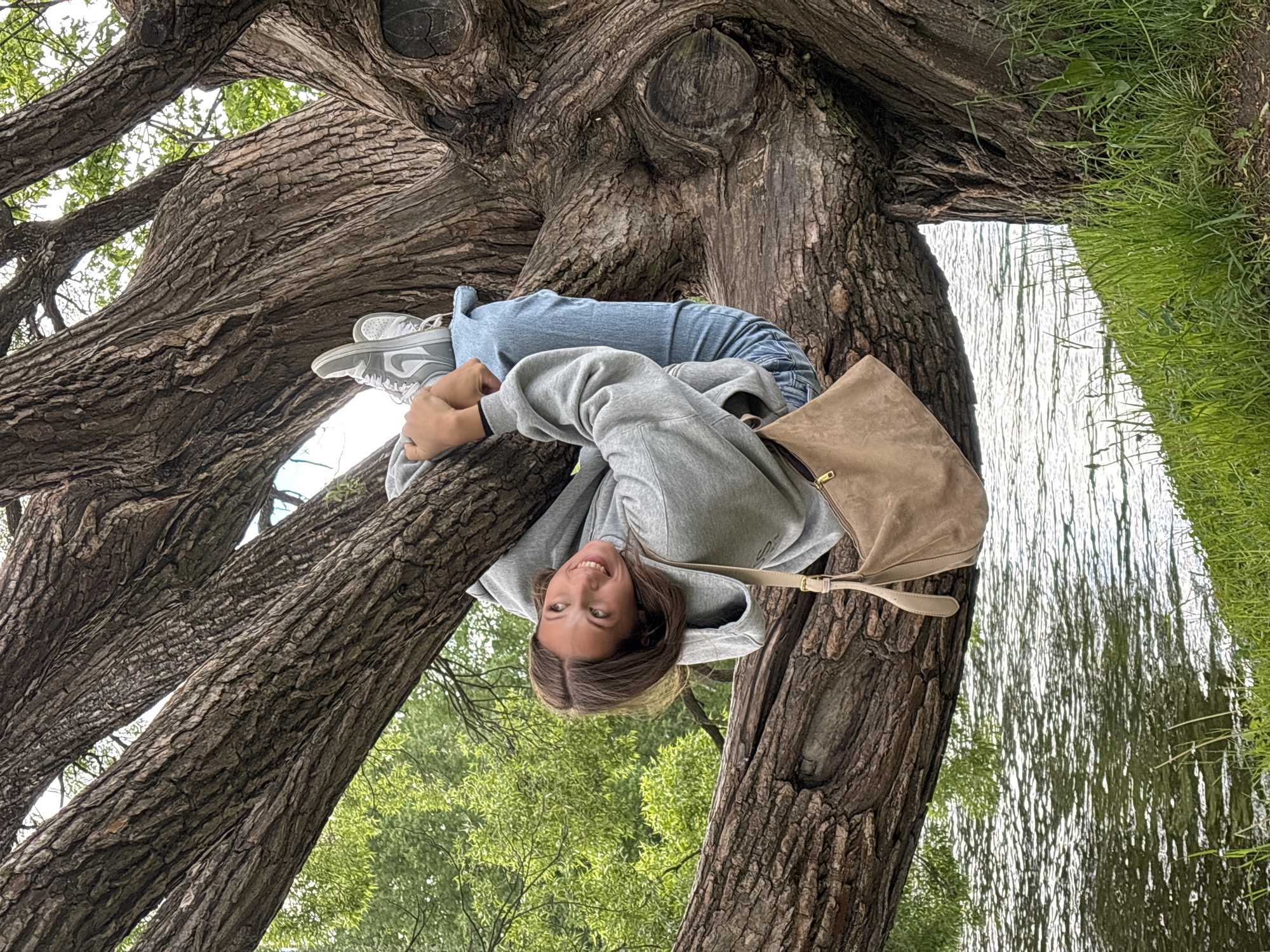 A person wearing a gray hoodie, blue jeans, and gray sneakers is sitting in a tree. The person is positioned between two thick branches, with their head and torso resting on one branch and their legs on the other. They are smiling and looking at the camera. A tan suede bag is slung over their shoulder. Behind the tree, water and grass are visible, indicating a waterfront location. Based on the trees, water and flora, the location of this image is likely Saint Petersburg, Russia.