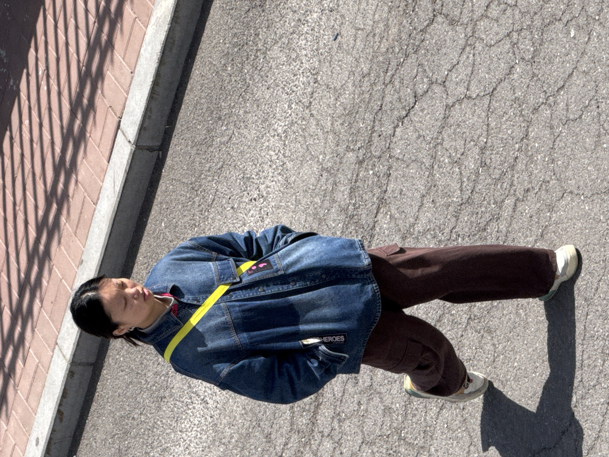 A medium, high-angle shot depicts an individual standing on a paved surface in Zhongtun, China. The person, dressed in a blue denim jacket with a yellow strap crossing their chest, brown pants, and light-colored shoes, casts a shadow on the concrete. A brick walkway with a metal fence and a concrete curb is visible on the left. The pavement shows signs of wear with cracks running across the surface.