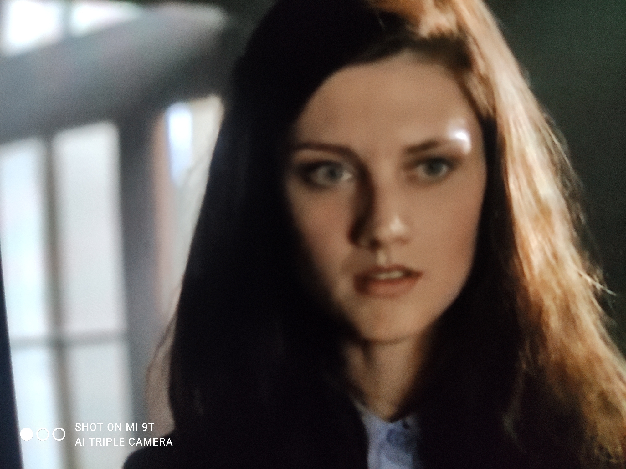 A young woman with long auburn hair and fair skin is pictured in a close-up, slightly blurred shot. She is looking towards the viewer with light blue eyes. Her lips are slightly parted, and she is wearing a light blue collared shirt. The background is dark and out of focus, with faint vertical lines that suggest a window or blinds. The lower left corner of the image displays a watermark that reads "SHOT ON MI 9T AI TRIPLE CAMERA." The specific location within Khazar Raion, Azerbaijan, cannot be determined from the visual information provided.