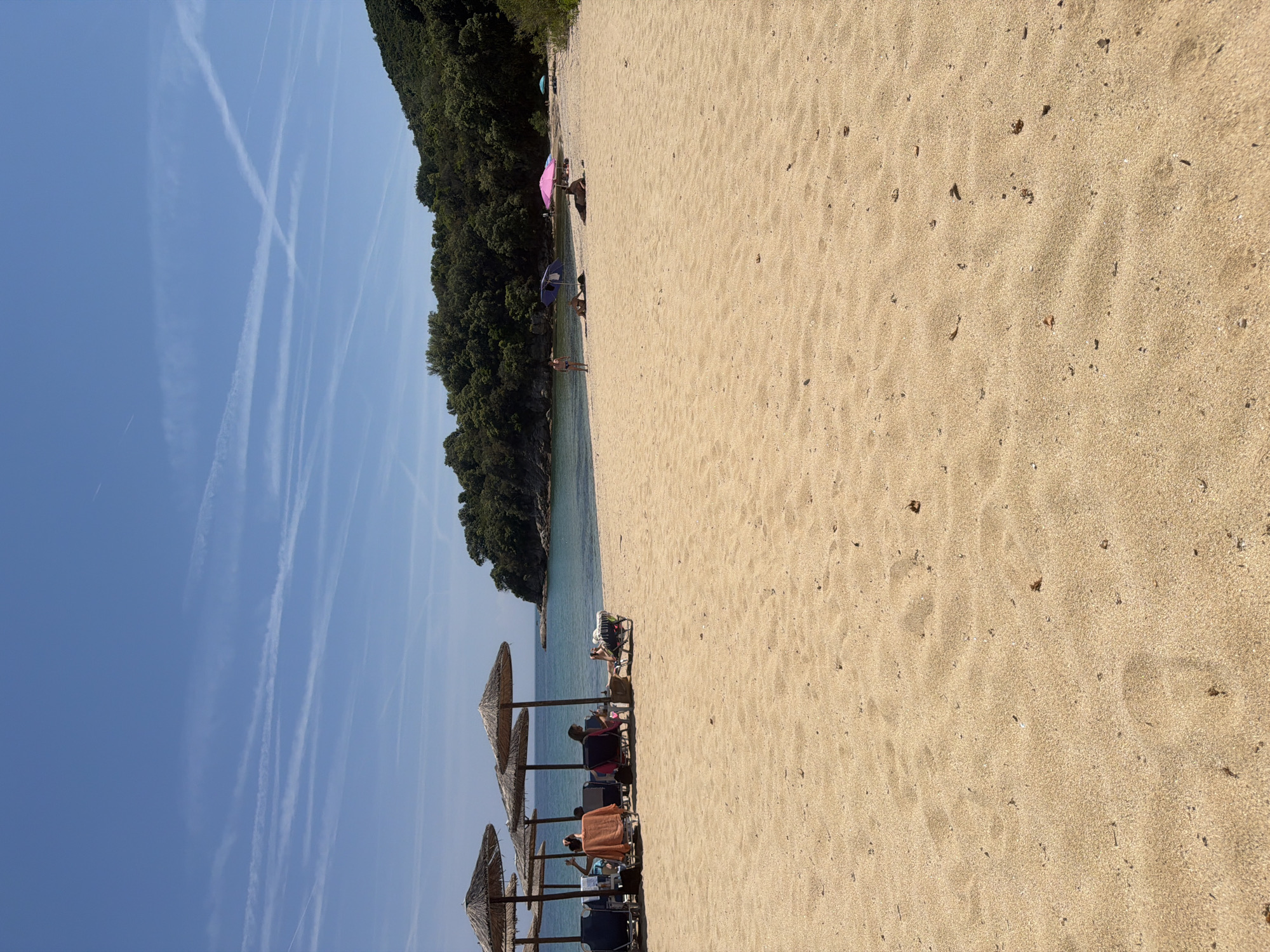 This image captures a wide-angle view of a sandy beach in Olympiada, Greece, under a clear, bright blue sky with numerous intersecting white aircraft contrails. The foreground is dominated by golden, fine-grained sand, showing subtle undulations, scattered small dark debris, and faint footprints.
In the midground, toward the left, a cluster of four thatched beach umbrellas is visible, each shading blue reclining beach chairs. Beneath these umbrellas, at least four individuals are present; one person wearing an orange/brown top is seen from behind, facing the sea, while others are partially obscured or also facing away. To the right of this group, further down the beach, multiple individuals are scattered across the sand. One person lies prone under a small blue umbrella, while another stands near the water's edge in a dark swimsuit. Further to the right, two people are situated under a bright pink umbrella, and another person is seen lying on the sand without an umbrella. A light blue umbrella is also visible in the distance.
The background features the calm, clear turquoise waters of the sea, which transitions to a deeper blue further from the shore. On the left side, a rocky headland covered in dense green Mediterranean vegetation extends into the water. The entire coastline behind the beach is flanked by lush, green, forested hills rising gently. The overall scene conveys a sunny, leisurely day at the beach.