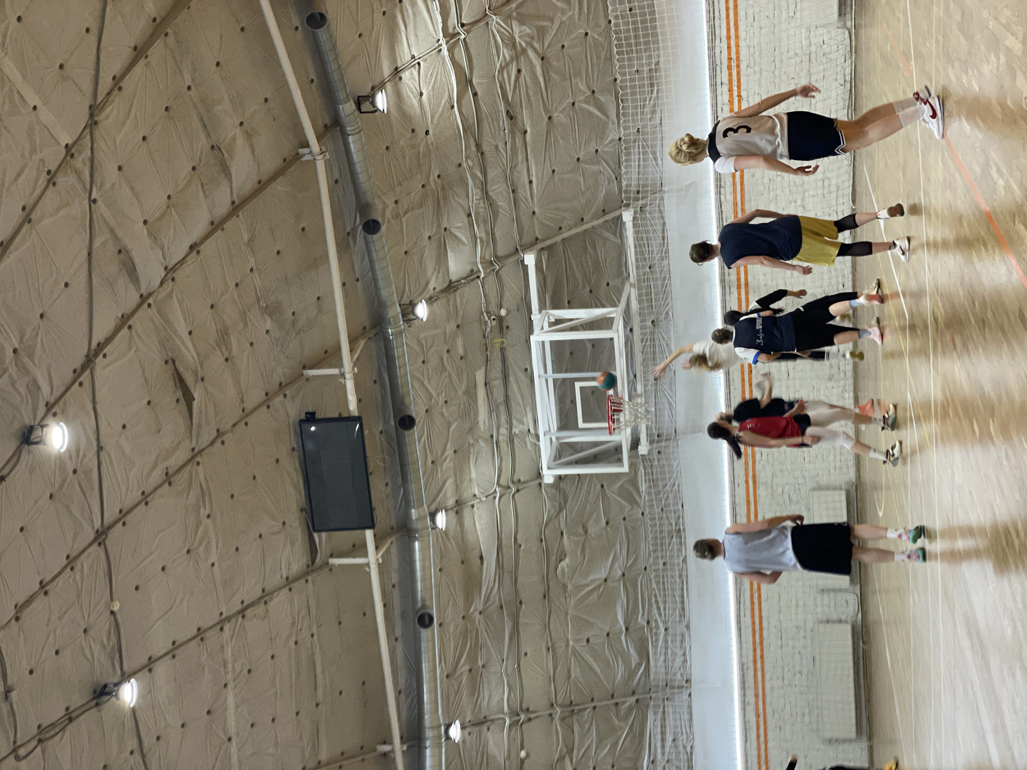 A group of people are playing basketball indoors in a gymnasium. The ceiling is covered in quilted, sound-dampening material, with overhead lighting and ductwork. There is a scoreboard mounted on the wall. The players are wearing athletic clothing, and some are in motion near the basketball hoop. The location is possibly in Saint Petersburg, Russia, based on the image's origins.