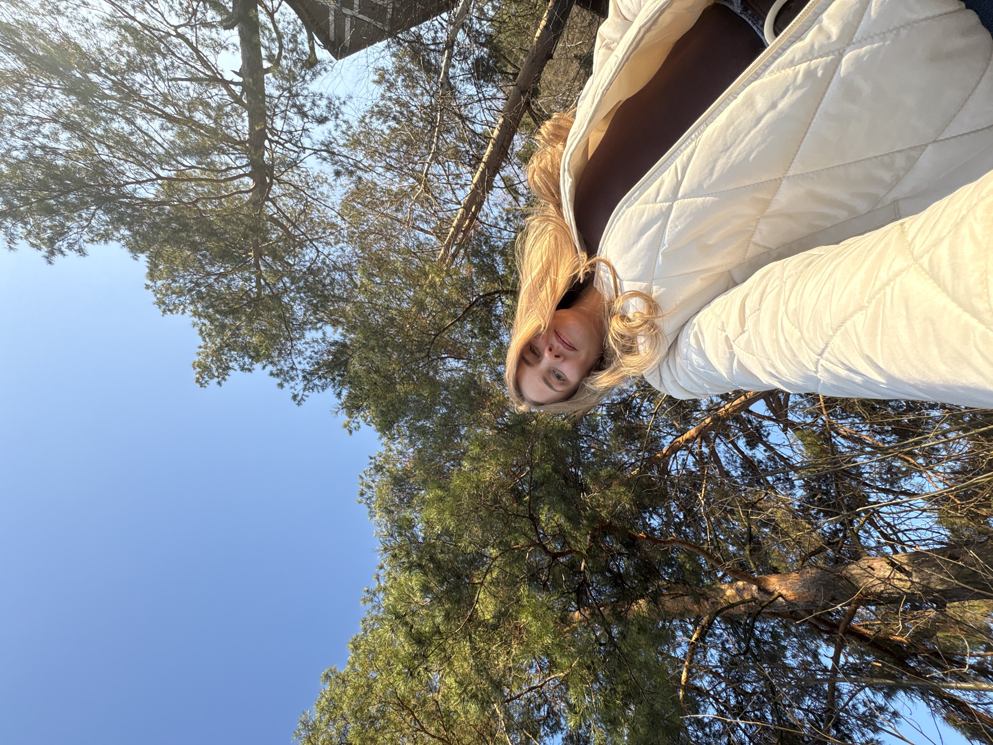 A woman with blonde hair is the primary subject of the image, wearing a brown top and a white quilted jacket. The background shows a clear blue sky and a dense cluster of pine trees. The perspective is from a low angle, looking up. The location appears to be a wooded area, possibly in Chaikovsky, Russia.