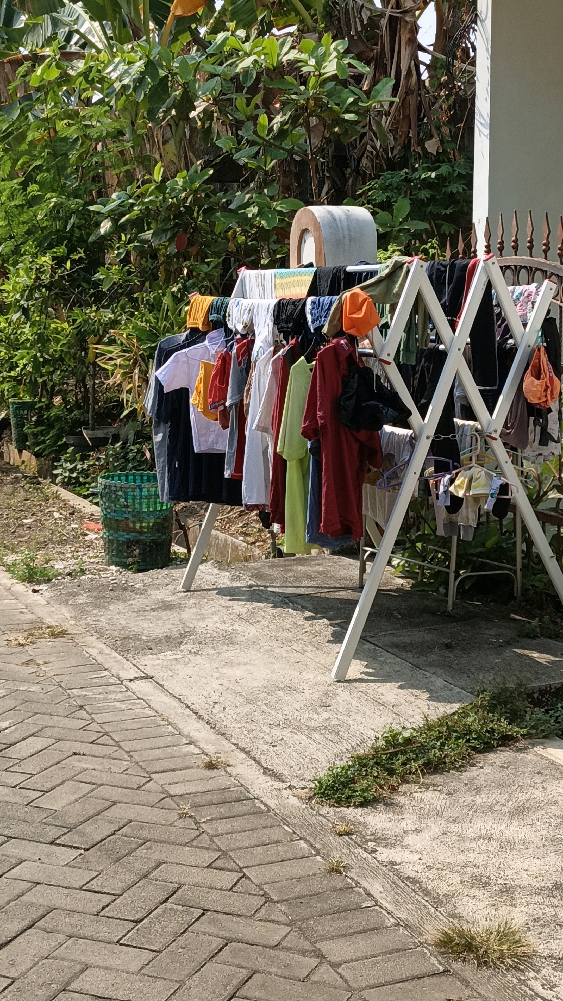 This image depicts an outdoor clothesline with a variety of clothing items hanging on it. The clothesline is a white, collapsible A-frame style rack. Among the visible clothing items are t-shirts, pants, and what appear to be undergarments, in various colors including white, black, red, yellow, green, and blue. Some items are on hangers, while others are clipped directly to the lines.
To the left of the clothesline, there is a green, woven plastic basket. The ground area is a mixture of concrete paving stones laid in a herringbone pattern and a rough concrete surface. Greenery, including bushes and trees with large leaves, forms a dense backdrop behind the clothesline. A rustic metal fence is visible behind the clothesline, and part of a building's white wall is seen on the far right. The scene is bathed in sunlight, casting shadows.
Based on visual cues, the location appears to be an outdoor residential area in a tropical climate, consistent with Cikal, Indonesia. The presence of lush vegetation and the style of the outdoor setting are indicative of this region. There are no people visible in the image.