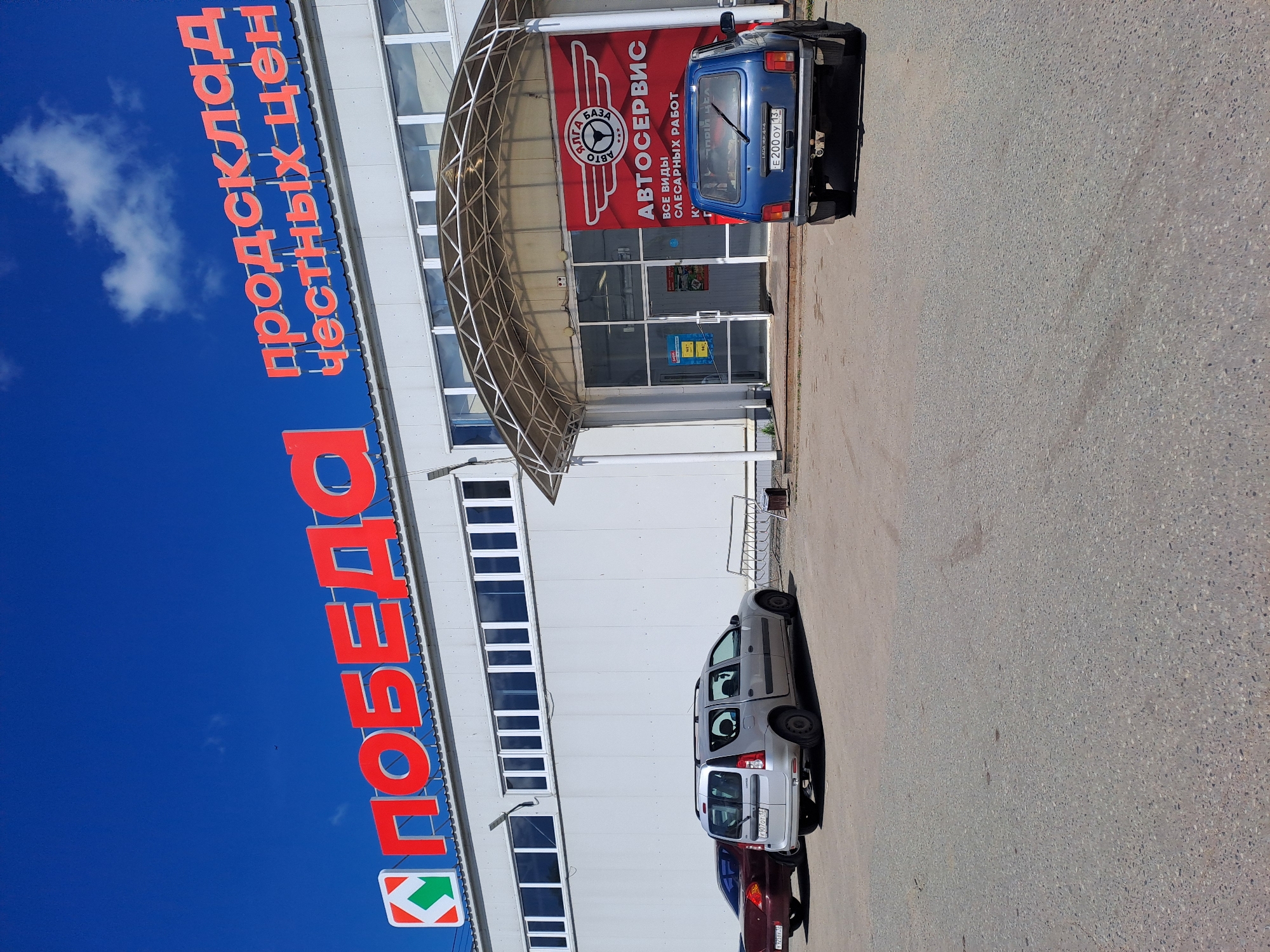 A daytime outdoor scene in Yalga, Russia, features a commercial building complex under a clear blue sky with sparse white clouds. The main building, tall and white with numerous vertical rectangular windows, has large red Cyrillic text "ПОБЕДА" (Pobeda - Victory) and "продсклад честных цен" (Honest Price Food Warehouse) affixed to its side, indicating a store or warehouse.
In front of the building, there's a paved area serving as a parking lot. Two cars are prominently visible. A silver station wagon, possibly a Lada Largus, is parked with its rear facing the viewer. The text "LADA" and "Largus" are visible on its tailgate, and its license plate partially shows numbers and ends with "116". Behind it, part of a maroon or dark red car is visible. To the right, a blue Lada Niva (or similar model) is parked, its rear also facing the viewer, displaying a license plate with the registration "E200OY 13".
An entrance to what appears to be an auto service center is located to the right of the main building. It features a translucent arched canopy over glass doors. A large red banner above and around the entrance proudly displays "АВТОСЕРВИС" (Avtoservis - Car Service) and "АВТО БАЗА" (Avto Baza - Auto Base), accompanied by a logo resembling a steering wheel. Below these, the text "ВСЕ ВИДЫ СЛЕСАРНЫХ РАБОТ" (All types of repair work) clarifies the services offered. No people are visible, and the scene is calm, suggesting a typical business day.