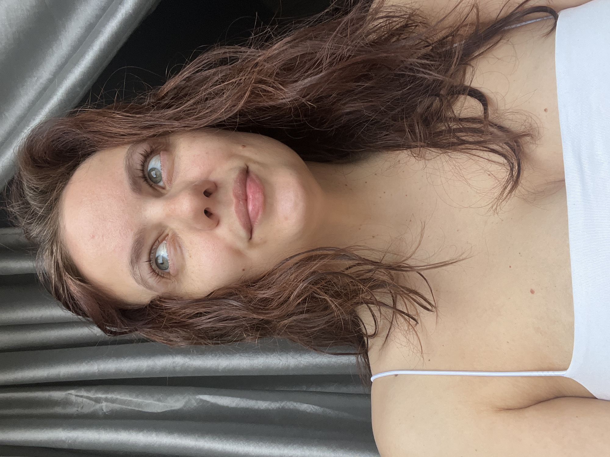 A selfie, taken indoors, features a young woman with brown hair, green eyes, and fair skin. She is wearing a white tank top with thin straps. The background consists of a textured grey curtain. There is no information in the image to connect it to the city of Chaikovsky, Russia.