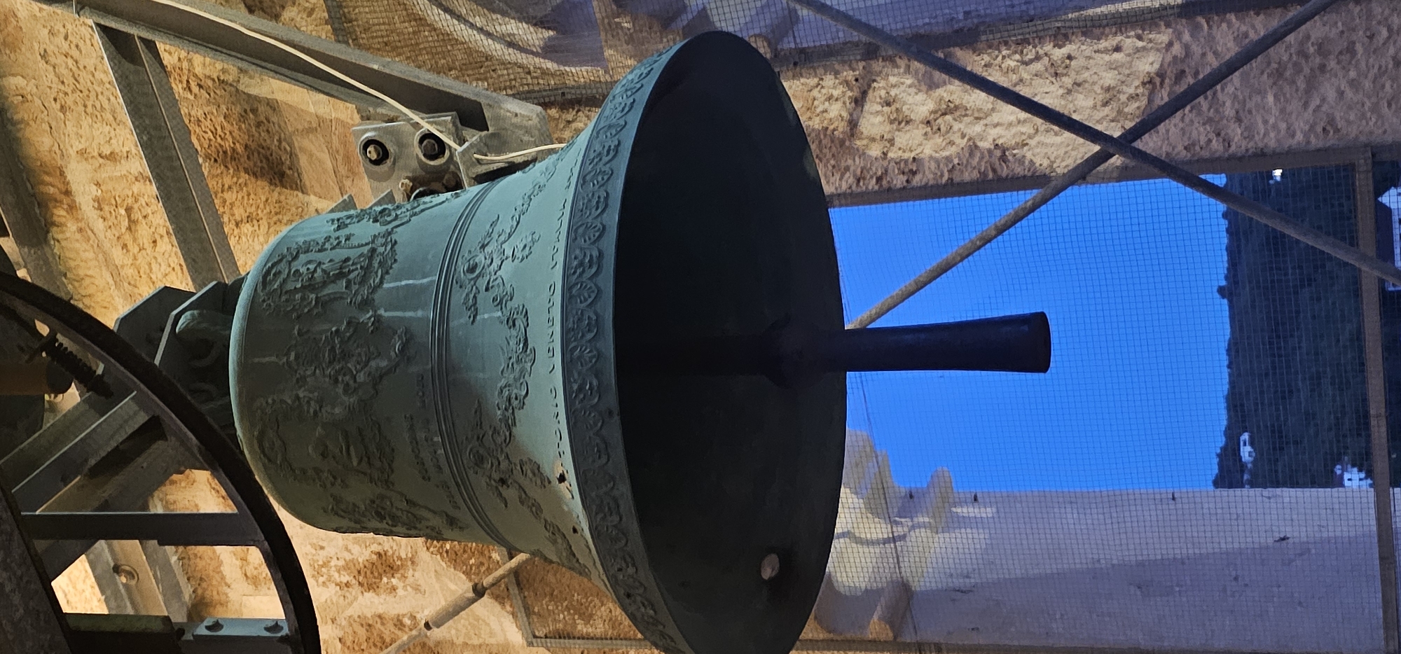 The image shows a close-up, low-angle view of a large, green bell hanging within a stone structure, likely a bell tower or belfry. The bell has ornate carvings and inscriptions, including text that appears to be "VITTORIO VENETO ITALIA". The bell is suspended by a dark, metal rod. Behind the bell, a bright blue sky is visible through a mesh screen. In the distance, a hilly landscape with scattered white buildings can be seen, illuminated by what appears to be dusk or twilight. The stone walls of the structure are rough and textured. Structural metal beams and supports are also visible, some appearing rusted. The overall scene suggests an outdoor setting, likely a historical or religious site. The inscription mentioning "Vittorio Veneto Italia" strongly suggests the bell was made in or for Italy, and its presence in Grad Supetar, Croatia, would imply a history of trade or connection between the two regions.