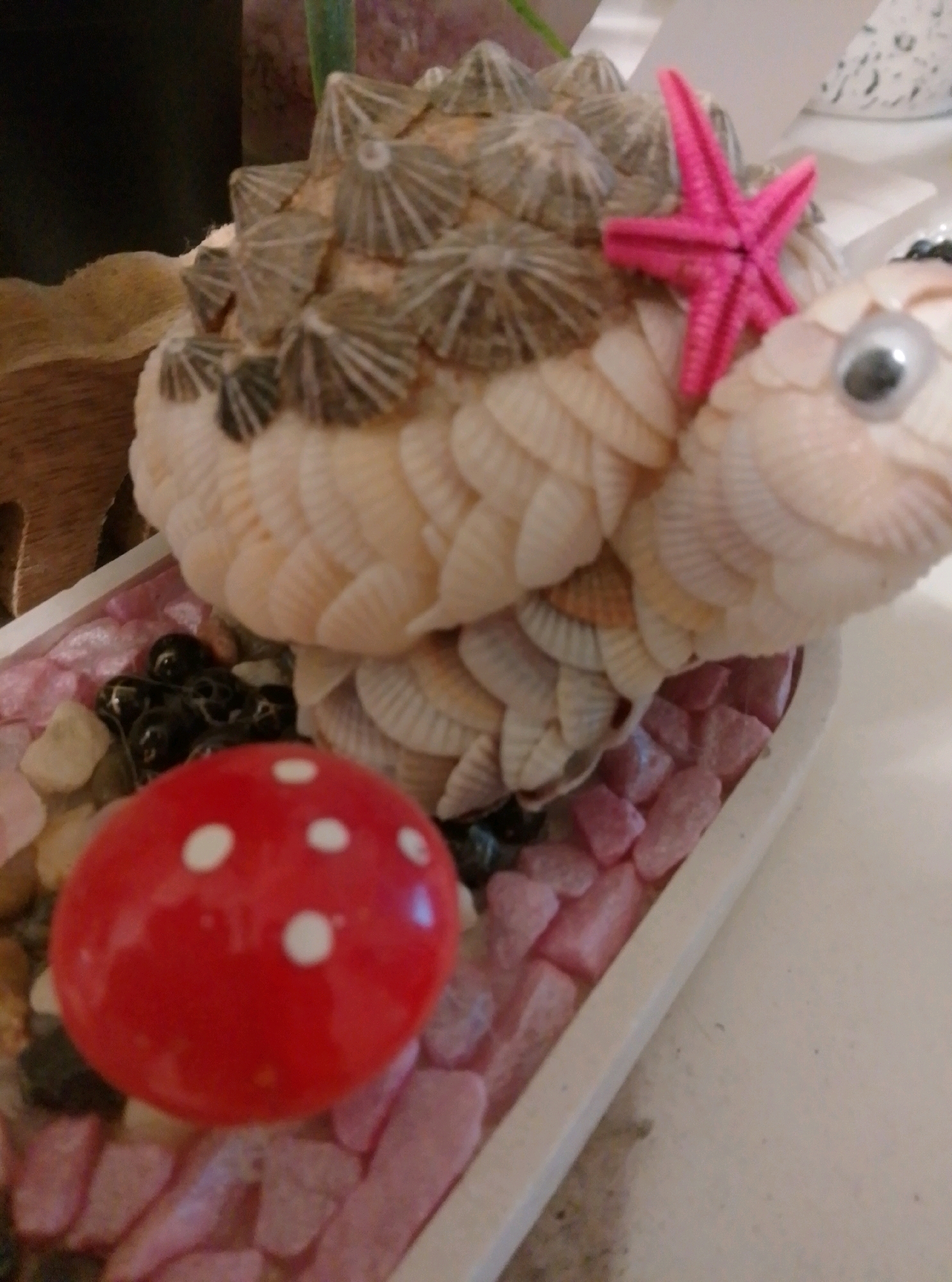 This is a close-up, slightly angled, indoor photograph of a decorative seashell fish. The fish is crafted from numerous small, pale, overlapping seashells, forming its body and fins. Its back is covered in what appear to be barnacle shells, and it has a googly eye on its right side. A small, bright pink, plastic starfish is affixed to its dorsal fin. The fish rests in a shallow, white rectangular container filled with small, pink and clear gravel. In the foreground, a small, bright red, round object with white polka dots, resembling a mushroom, is placed amongst the gravel. The background is dark and out of focus, with a hint of greenery and a white patterned object visible on the right. The scene does not depict any specific location in Wedel, Germany.