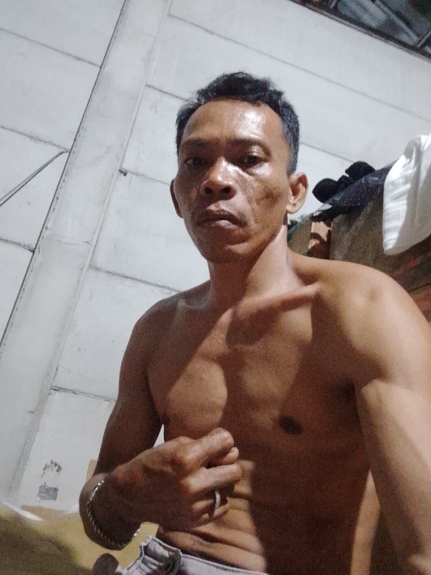 The image shows a shirtless man in Bukit Ketok, Indonesia. The man has short, dark hair and a dark complexion. He is looking directly at the camera with a serious expression. He is wearing a silver bracelet on his left wrist and a ring on his left hand. The background is a wall with a concrete panel design. There is also a pile of clothes or fabric in the upper right corner.