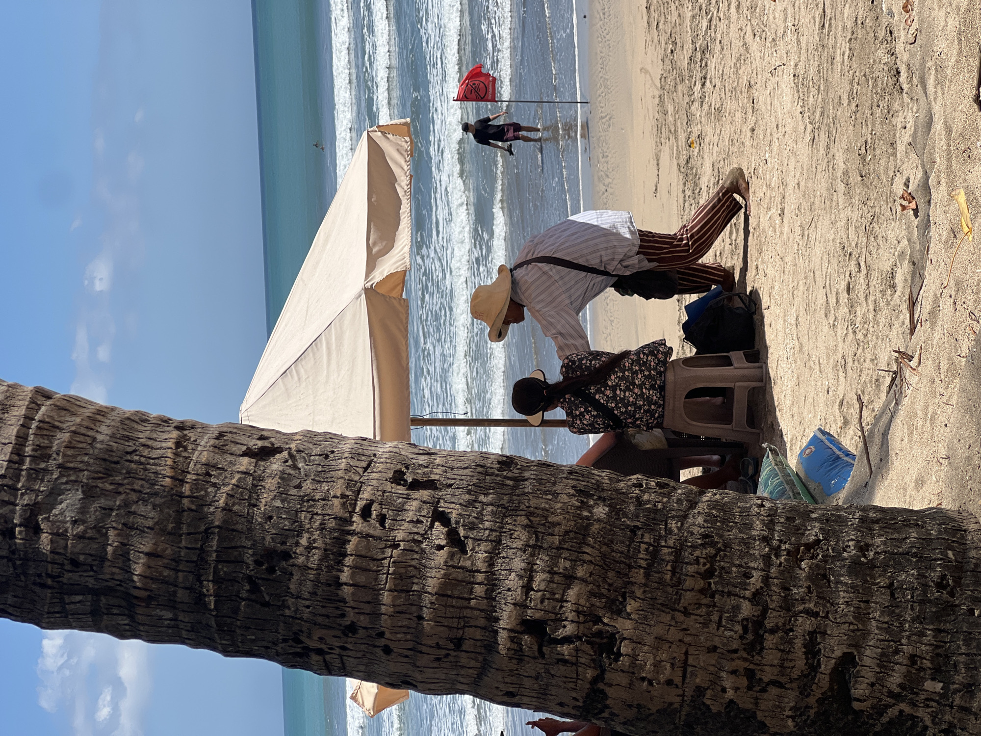 In Kuta, Indonesia, a beach scene unfolds with a large palm tree trunk dominating the foreground on the left. Behind it, under a light-colored beach umbrella, two individuals are seated. One, wearing a floral-patterned top and a dark visor, sits on a light-colored stool. The other, wearing a straw hat, a striped shirt, and striped trousers, leans forward and appears to be interacting with the seated person. A dark bag rests on the sand near them. In the middle ground, the ocean presents gentle waves. A man in a dark t-shirt and shorts walks in the shallow water, carrying flip-flops, near a red flag planted in the sand. The sky is a clear blue with a few wispy clouds. The beach is sandy, with some dry leaves and debris scattered on the ground.