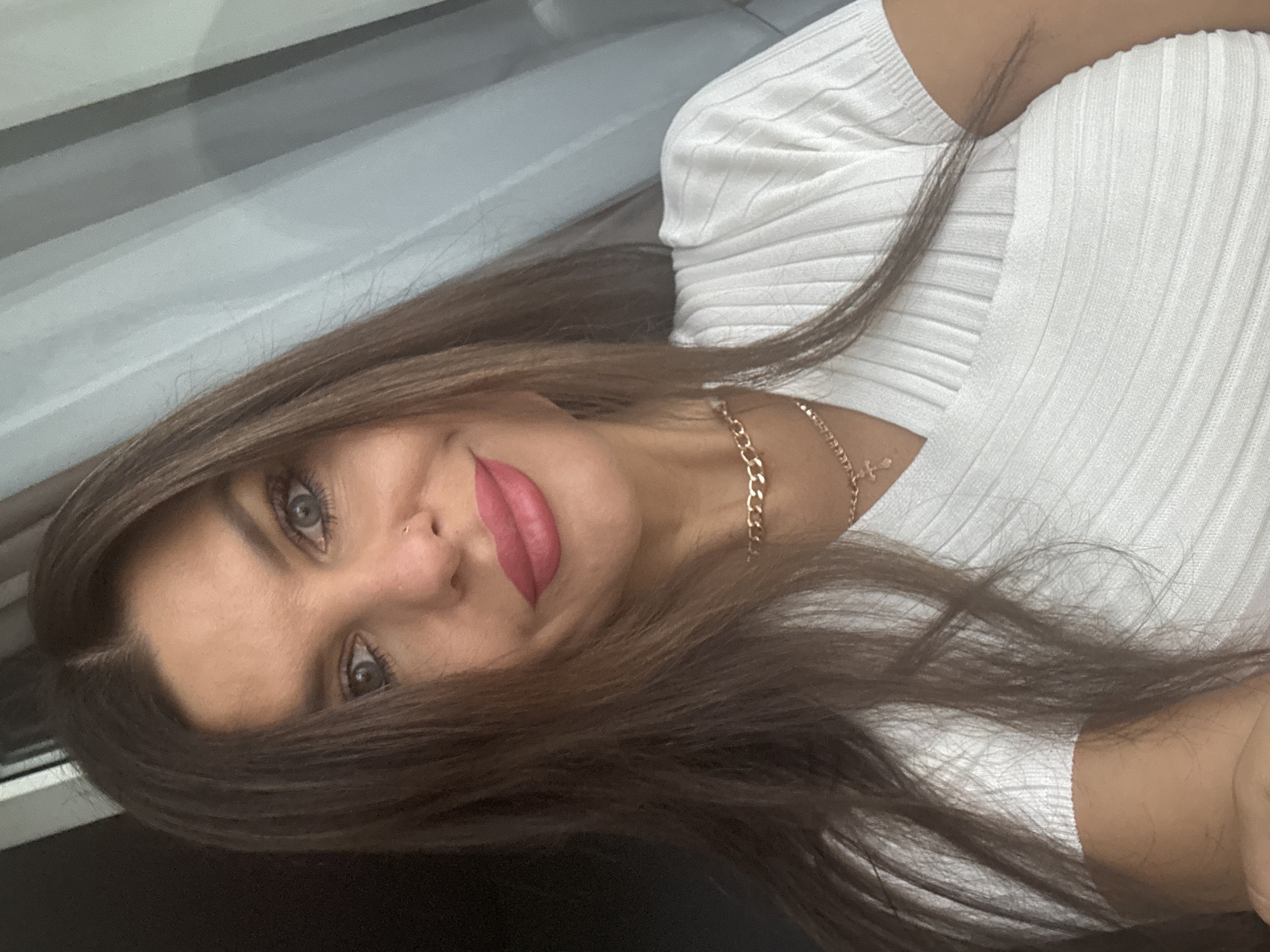 A medium close-up selfie shows a woman with long, dark brown hair, blue eyes, and full, pink lips. She is wearing a white, ribbed top and a gold chain necklace with a cross pendant. The background is blurred, showing vertical lines that suggest curtains or blinds. The lighting is soft and natural. The image does not contain any information to indicate the location in the city of Chaikovsky, Russia.