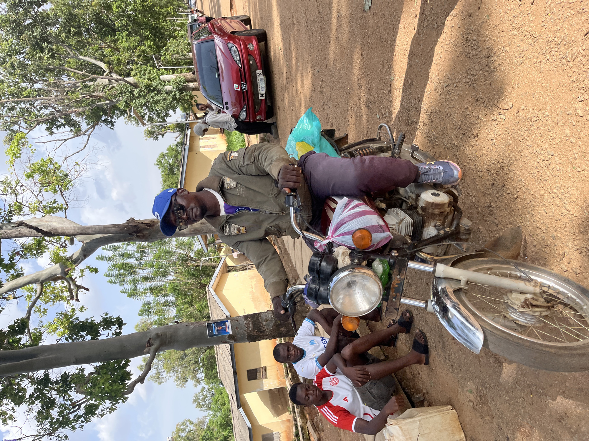 The image depicts a scene on a dirt road in Jos, Nigeria, where a man wearing a blue baseball cap and sunglasses is seated on a motorcycle. He is dressed in a brown jacket and dark pants. To his left, two young men are sitting on the ground. The man on the left wears a white and red soccer jersey, and the man to his right wears a white jersey with blue accents. Behind the motorcycle, a red Peugeot car is parked on the side of the road, with a few other individuals visible in the background near a building. The environment appears to be a residential area with houses and trees. The motorcycle has a bag with a blue tarp on the back and a striped package tied to the front. The ground is covered in dirt and small stones.