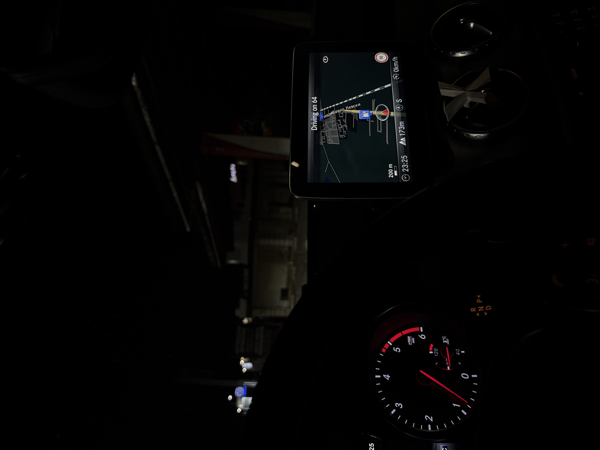 The image is taken from the interior of a car at night, likely in Plovdiv, Bulgaria, given the context of the navigation display showing Bulgarian street names. In the foreground, a tachometer is visible with a red needle pointing between 0 and 1, indicating a low engine RPM. The dashboard also shows a gear selector illuminated with R, N, P, and D.
A central infotainment screen displays a navigation interface. The screen indicates "Driving on 64" and shows a route with street names in Bulgarian, including "Иван Василевски" and "Левски". The current time is 23:25, and the car is estimated to be 173 meters from the next turn. The speed is displayed as 0 km/h, suggesting the car is stationary or moving very slowly. A fuel station icon is present on the map.
In the background, the exterior environment is dark, with illuminated buildings and streetlights visible through the car's windshield, consistent with an urban setting at night.