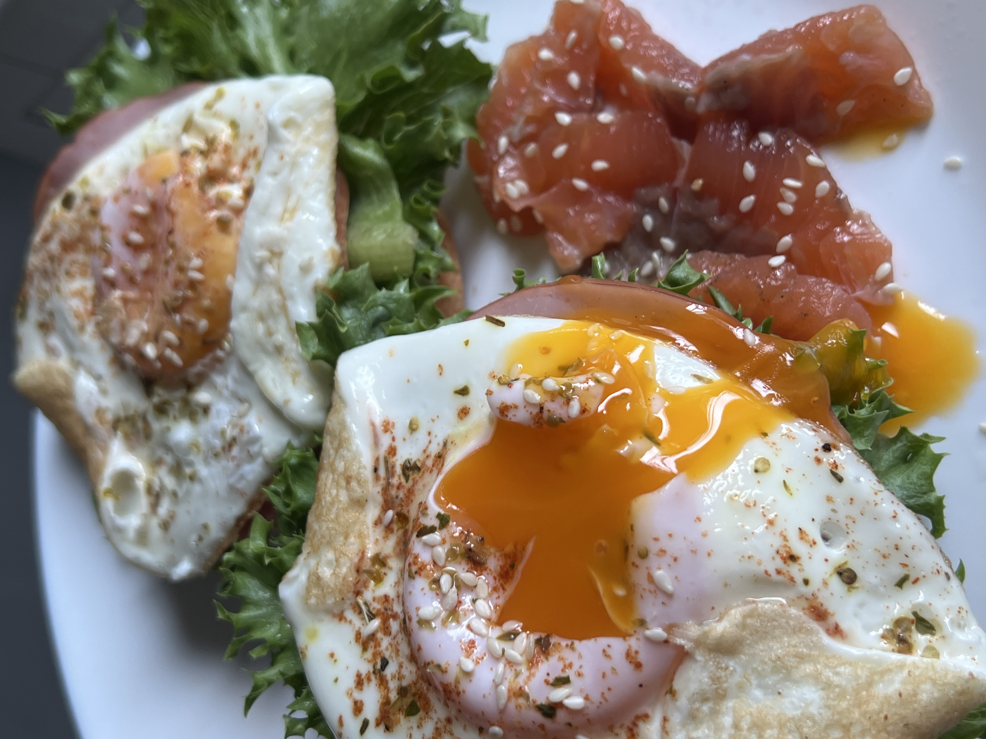 Two open-faced sandwiches with fried eggs and lettuce are presented on a white plate. One sandwich in the foreground features a sunny-side-up egg with a runny yolk, sprinkled with sesame seeds and red pepper flakes. The other sandwich, slightly behind, also has a fried egg and appears to be on a bed of lettuce and tomato. To the right of the sandwiches is a portion of what appears to be raw salmon, also sprinkled with sesame seeds. The image is a close-up shot taken from a high angle, focusing on the food. The location is not specified in the image.