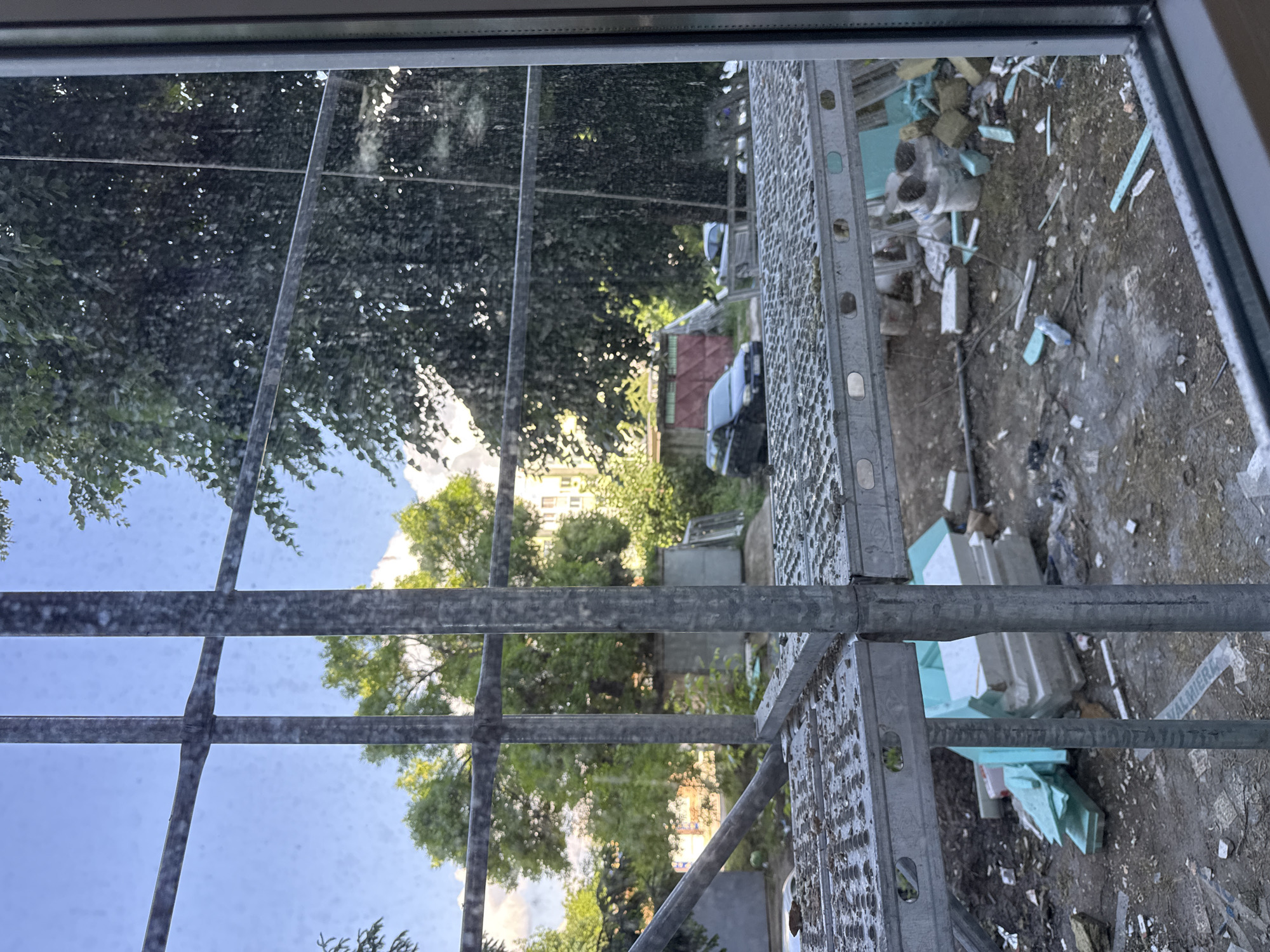 The image depicts a view from a window with a metal frame, looking out onto an outdoor scene in the city of Lovech, Bulgaria. A metal scaffolding structure partially obscures the view to the right. Beyond the scaffolding, there is a pile of debris, including pieces of white and blue foam. In the background, trees and a building can be seen, with a portion of a red-roofed structure visible. A vehicle is also present in the scene. The sky is visible through the trees. The photograph appears to have been taken during daylight hours.