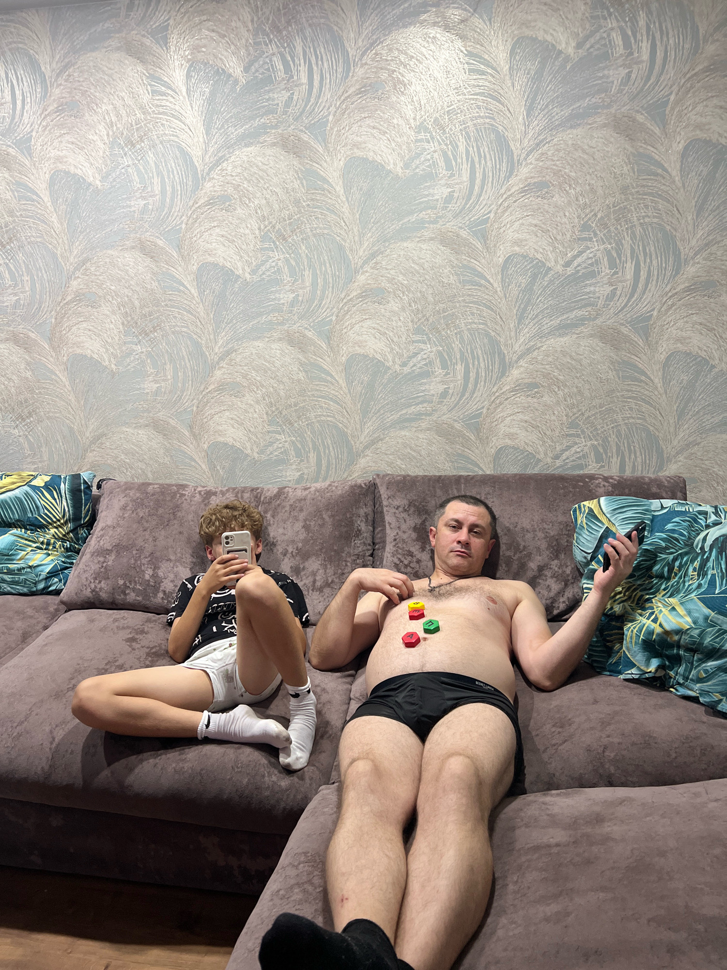The image depicts an indoor scene, likely a living room, featuring a large, taupe-colored sectional sofa. On the sofa, two individuals are present: an adult male lying down and a younger male sitting up. The adult male, bare-chested and wearing black underwear, is lying on his back with his head resting on a cushion. Three colorful, hexagonal objects (red, yellow, and green) are placed on his chest. He is holding a black mobile phone in his right hand.
The younger male, seated with his legs crossed, is wearing a black t-shirt with white graphics and white shorts. He is looking down at a white mobile phone he is holding in his hands. Both individuals are positioned on the same sofa, with the younger male to the left of the adult male. Decorative pillows with a tropical leaf pattern in blue, green, and yellow are visible on either side of the sofa.
The background wall is covered in patterned wallpaper, featuring a light blue base with a recurring textured design in a lighter, almost white, color, resembling stylized feathers or brushstrokes. The lighting in the room appears to be artificial, casting soft shadows. There is no indication of the specific location within Noviy, Russia, in the image itself.
