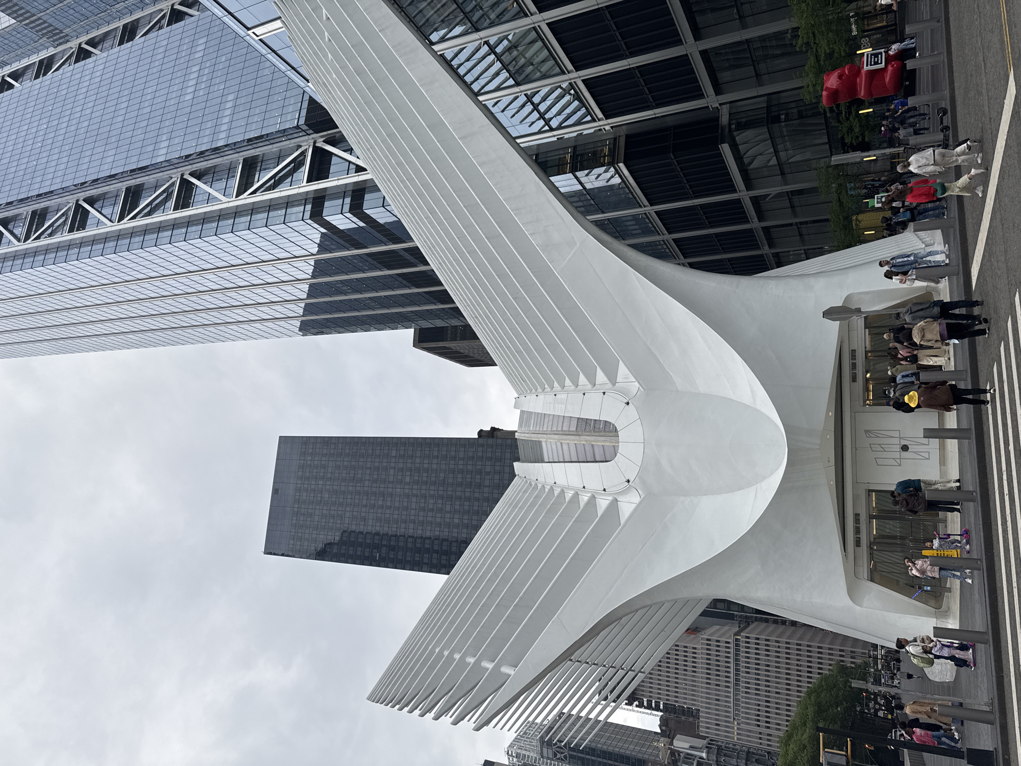 This photo shows the World Trade Center Transportation Hub, also known as the Oculus, in New York City. The Oculus is a white, wing-like structure. People are walking and standing around the entrance. Buildings are visible in the background. The sky is cloudy.