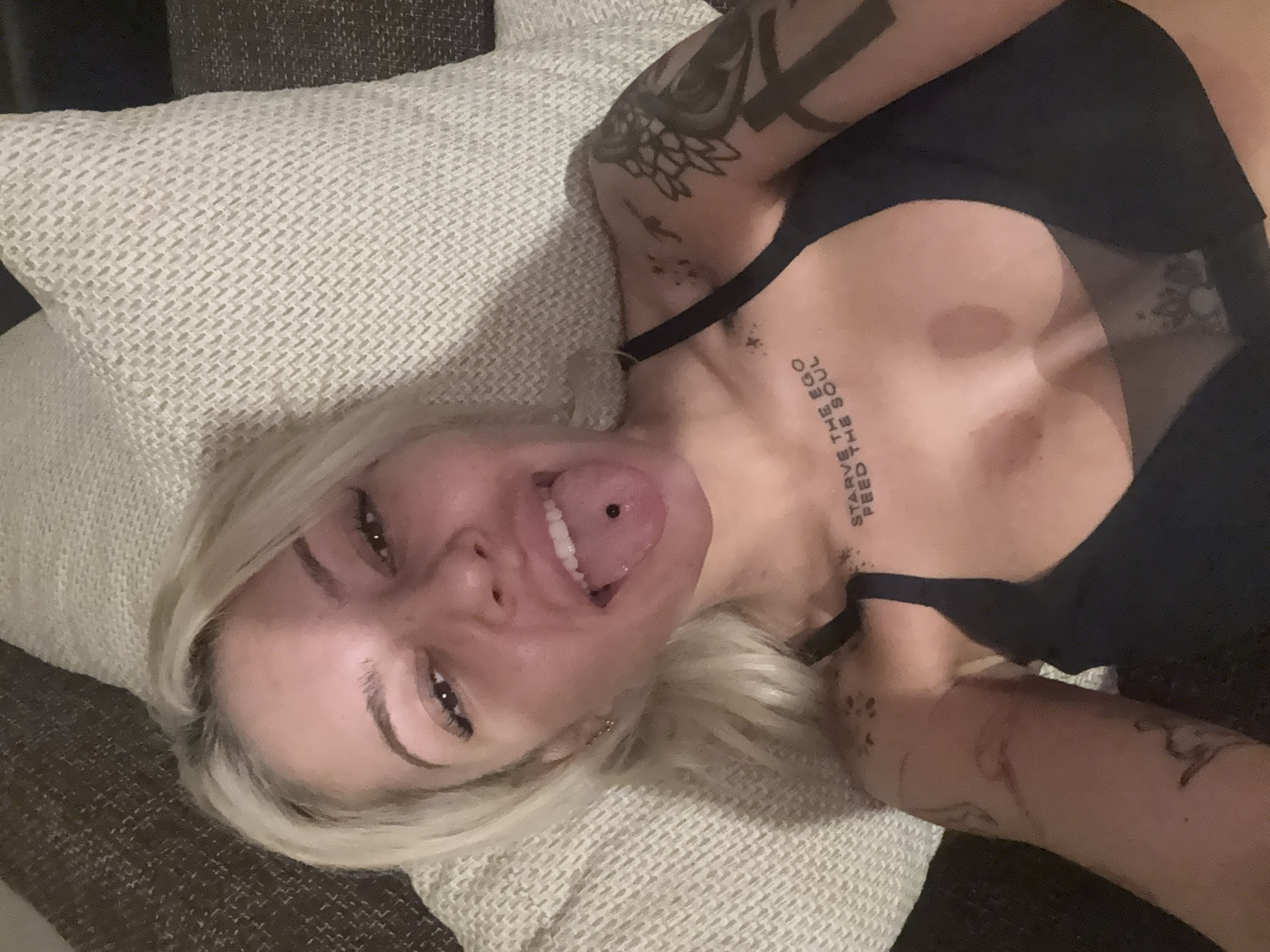 A blonde-haired person with multiple tattoos, including one on their chest that reads "Starve the Ego Feed the Soul," is lying on a textured, light-colored pillow on a dark-colored surface. The person is wearing a black bra or top. They have a pierced tongue and are sticking it out. The location is unknown.