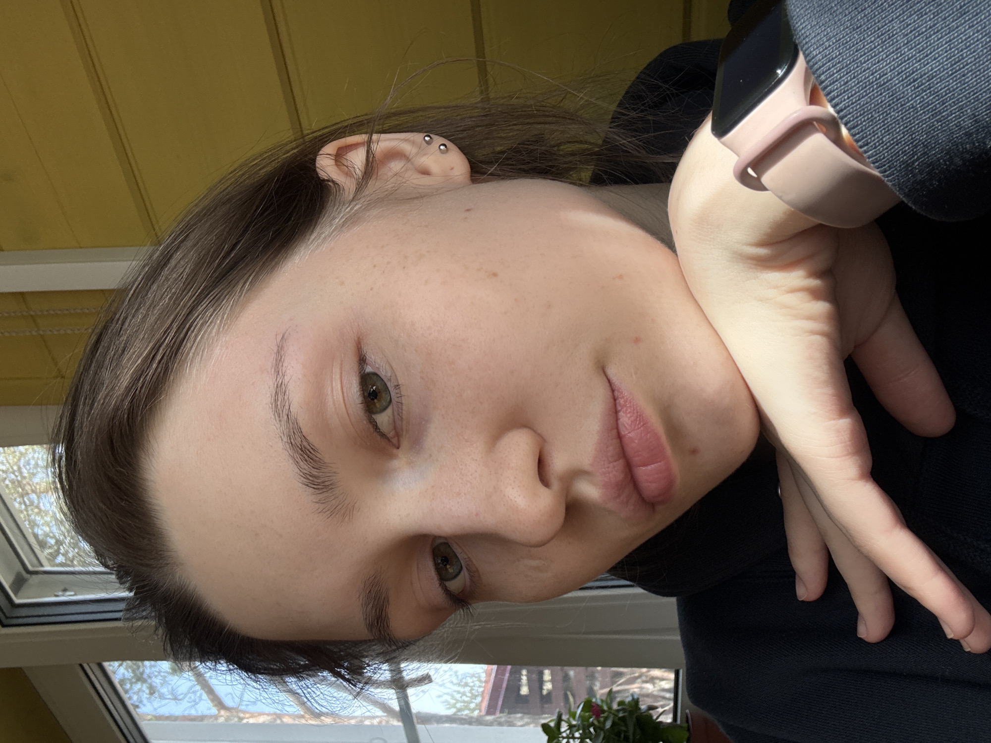 A close-up portrait of a young, light-skinned person with brown hair and green eyes. The person is wearing a dark hooded sweatshirt and a pink smartwatch on their left wrist. Their left hand rests on their cheek. The background includes a window with visible trees outside and a yellow wall.
I cannot determine if the picture was taken in the city of Ленинградская, Russia.