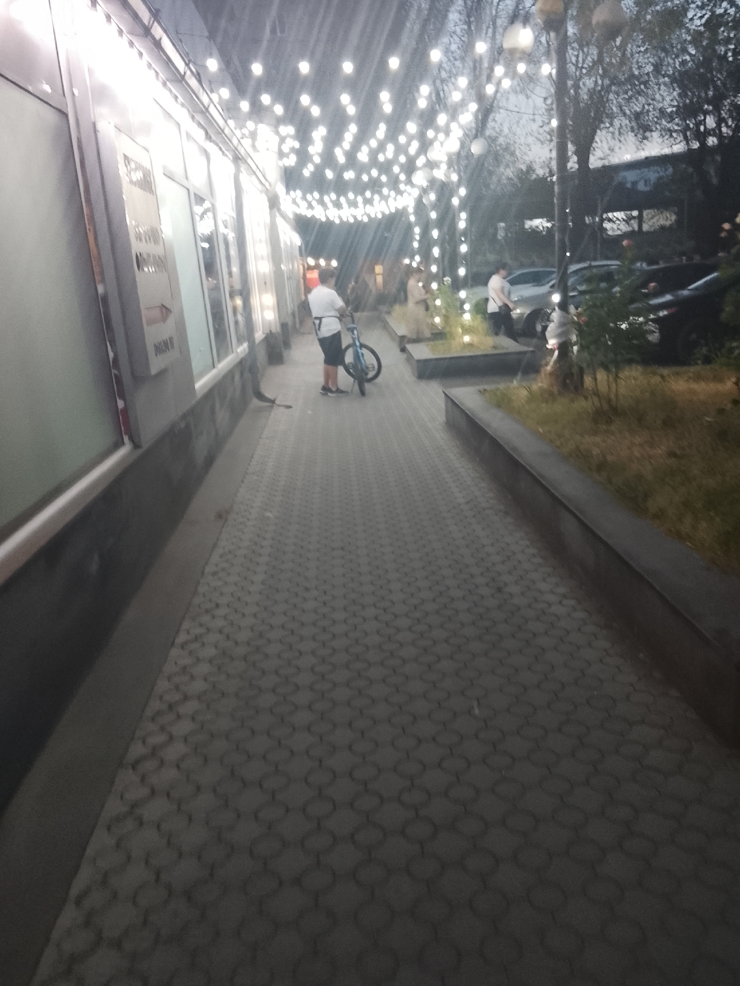 A full shot captures an outdoor scene in an urban setting, likely in the evening given the lighting. A paved walkway, composed of interlocking grey hexagonal tiles, stretches into the distance. To the left, a building facade features large glass windows and illuminated signage. In the middle ground, a person in a white t-shirt and dark shorts is seen standing with a blue bicycle. Further back, other individuals and parked cars are visible, along with trees and streetlights strung with numerous small, bright lights, creating a festive atmosphere. The overall environment suggests a public space, possibly a pedestrian zone or an area adjacent to shops or entertainment venues.