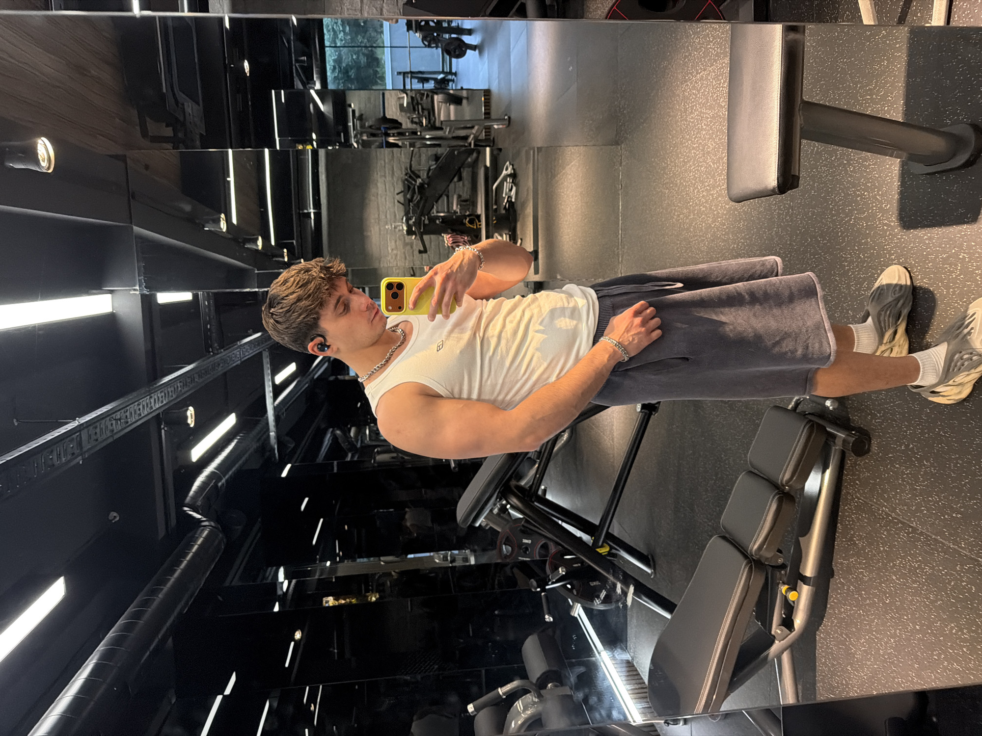A young man with brown hair is taking a selfie with a yellow cell phone in a gym. He is wearing a white tank top, grey shorts, white socks, and grey shoes. He also wears a chain necklace, bracelets, and what appears to be an earbud. He stands in front of a mirrored wall that reflects the gym interior, including various exercise machines. The gym features a dark interior with overhead lighting. The location is likely in a gym facility in Sofia, Bulgaria.
