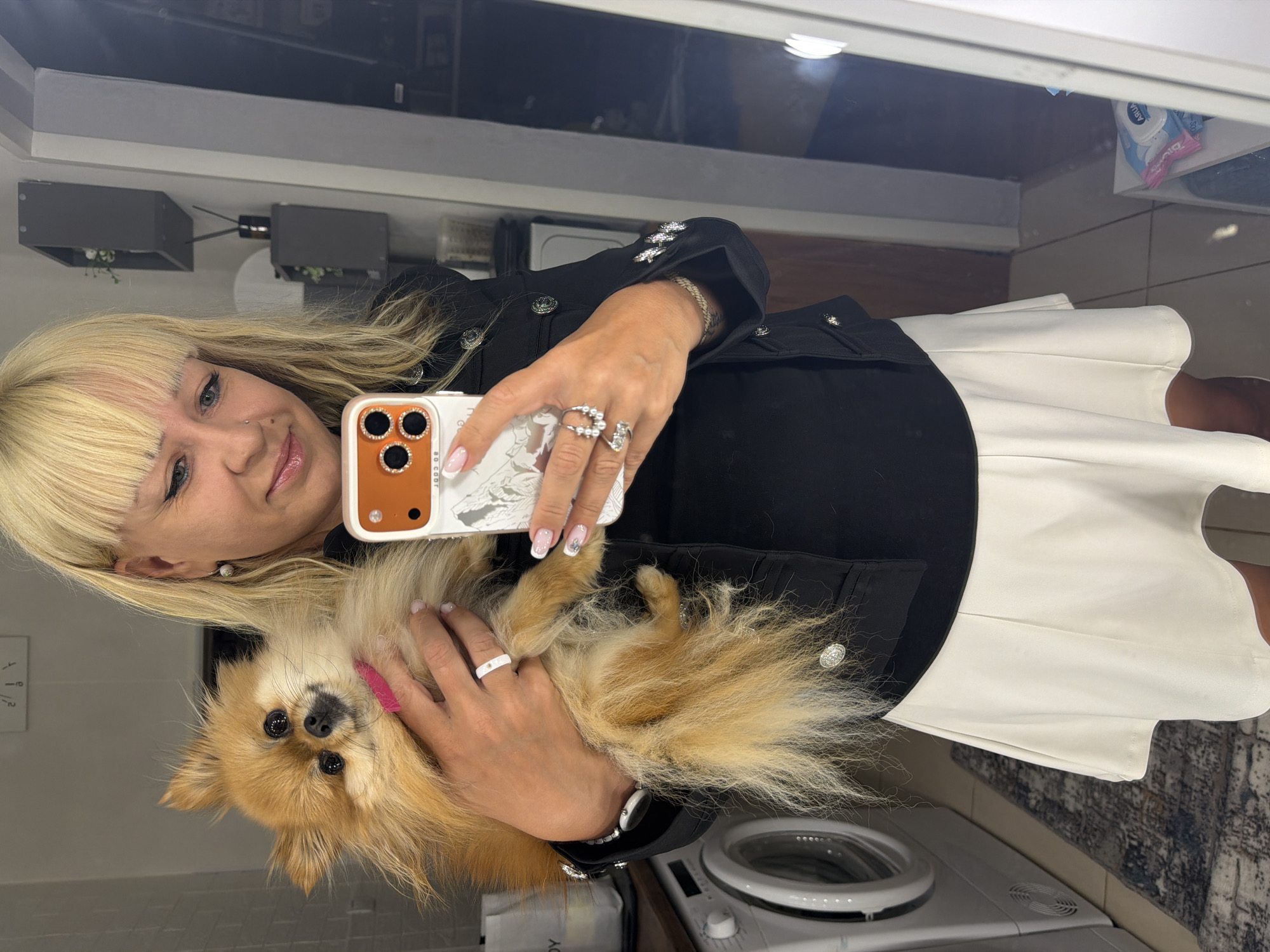 A woman with blonde hair is holding a small, tan-colored Pomeranian dog while taking a selfie. She is wearing a black jacket with silver buttons and a white skirt. The woman is wearing multiple rings and a watch. Behind her, there is a washing machine, a white tile wall, and a mirror reflecting the room. There is a box with text on the bottom left of the frame. The location is possibly a bathroom. The city of Chaikovsky, Russia cannot be confirmed from the visual data.
