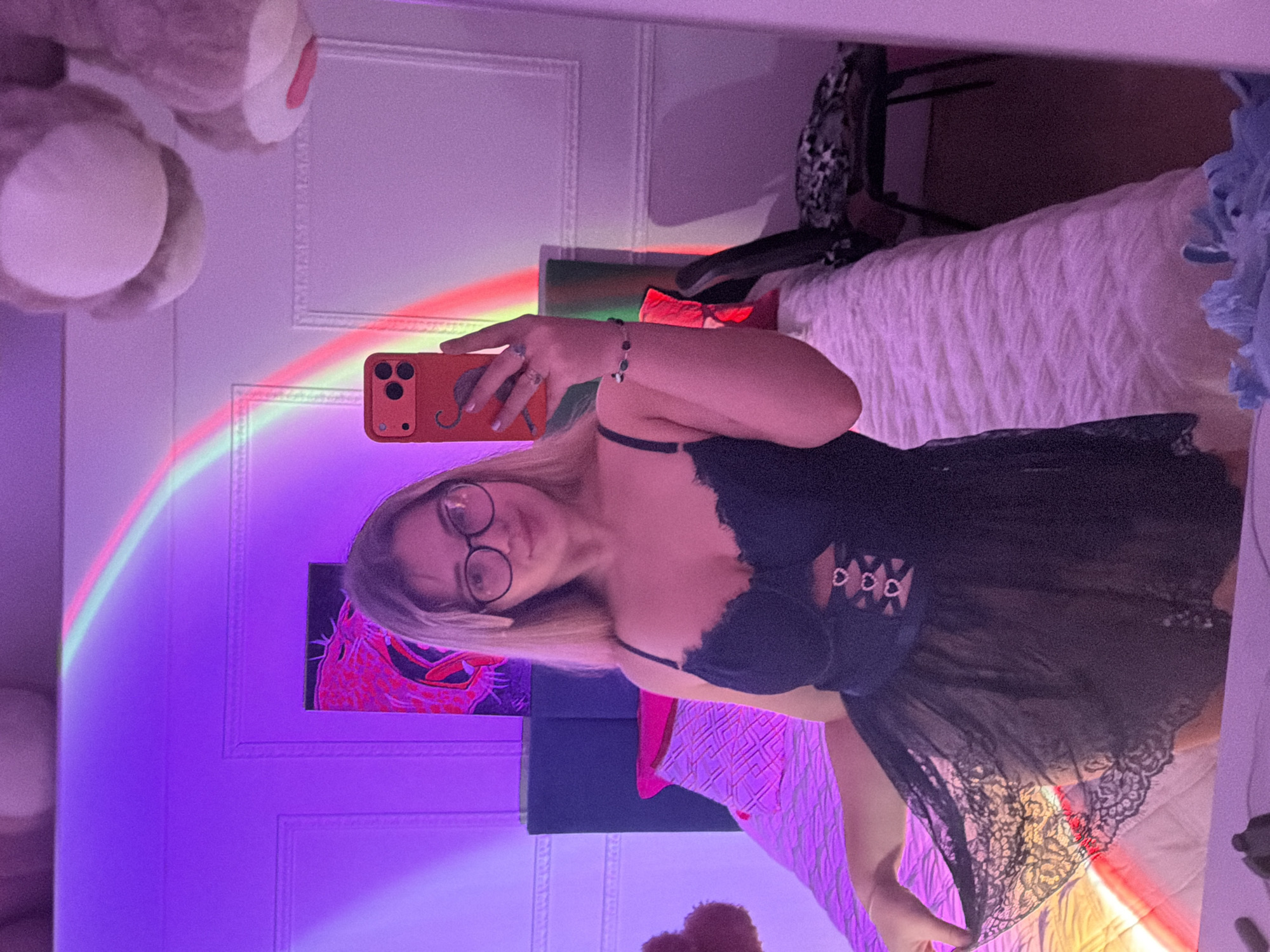 The image depicts a woman in a dimly lit room, seemingly taking a selfie. She is wearing a black negligee with lace trim. Her face is visible, and she is wearing glasses. She is holding an orange cell phone with a design on the back. The background includes a bed with a patterned bedspread, a teddy bear, and a light projecting a rainbow onto the wall. There is also a piece of art on the wall. It's difficult to confirm the exact location in Большая Шильна, Russia with only this image.