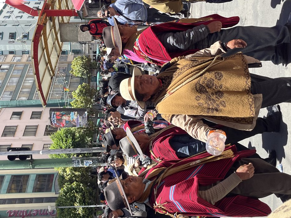 A crowd of people, many wearing traditional Andean attire, fills a city street. Men in hats and colorful ponchos stand out, some holding plastic bottles. Buildings line the background with trees and signs dotting the landscape. Overhead, a partial awning structure provides some shade. The scene captures a vibrant cultural gathering or procession.