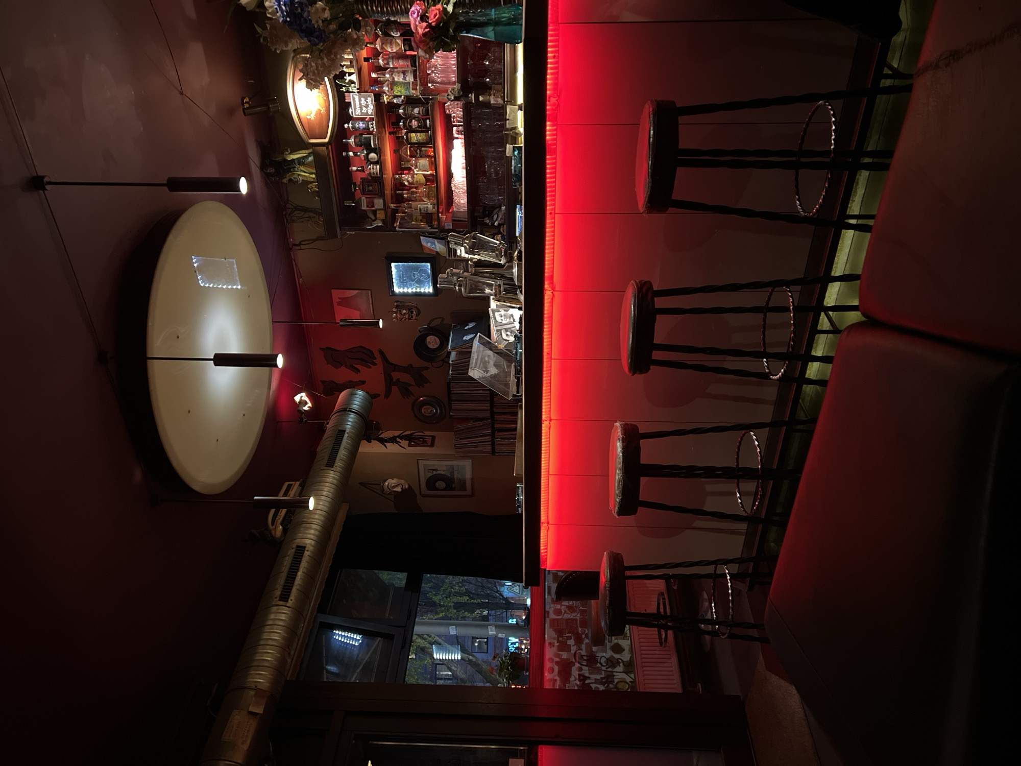 The photograph depicts an interior view of a bar, likely located in Berlin, Germany, judging by architectural styles and urban scenery visible through the windows.
The bar features a prominently illuminated counter, bathed in red light, with several high-backed stools positioned along it. A red sofa is situated on the right side of the photograph, adjacent to the stools. Behind the bar is a display of various bottles, glasses, and bar equipment, suggesting a well-stocked establishment. The walls are decorated with an eclectic mix of objects, including records and artwork. A large, round light fixture hangs on the wall to the left, further illuminating the scene. A corrugated metal pipe is visible. The environment appears dark and cozy. Outside the window is a view of urban buildings and trees. No people are visible in the shot.